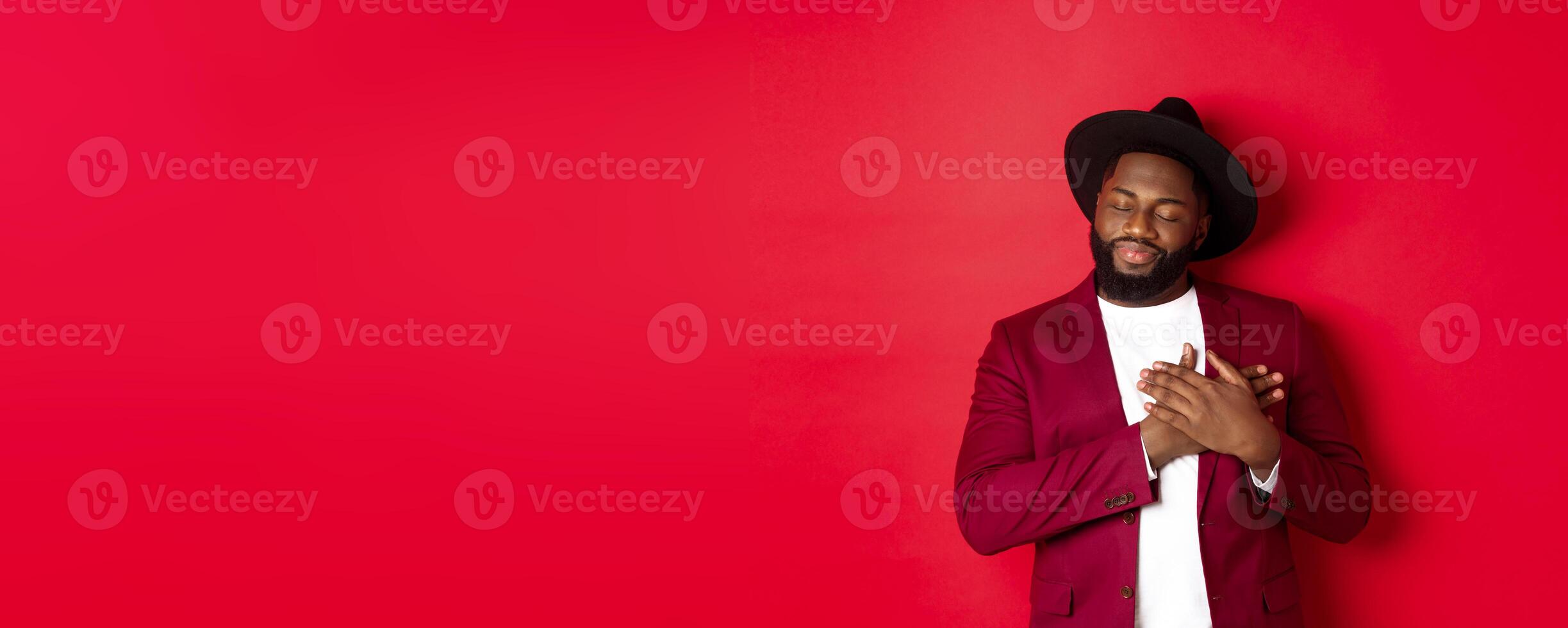 Fashion and party concept. Young bearded Black man in classy hat and jacket, holding hands on heart and smiling nostalgic, remember something, daydreaming against red background photo