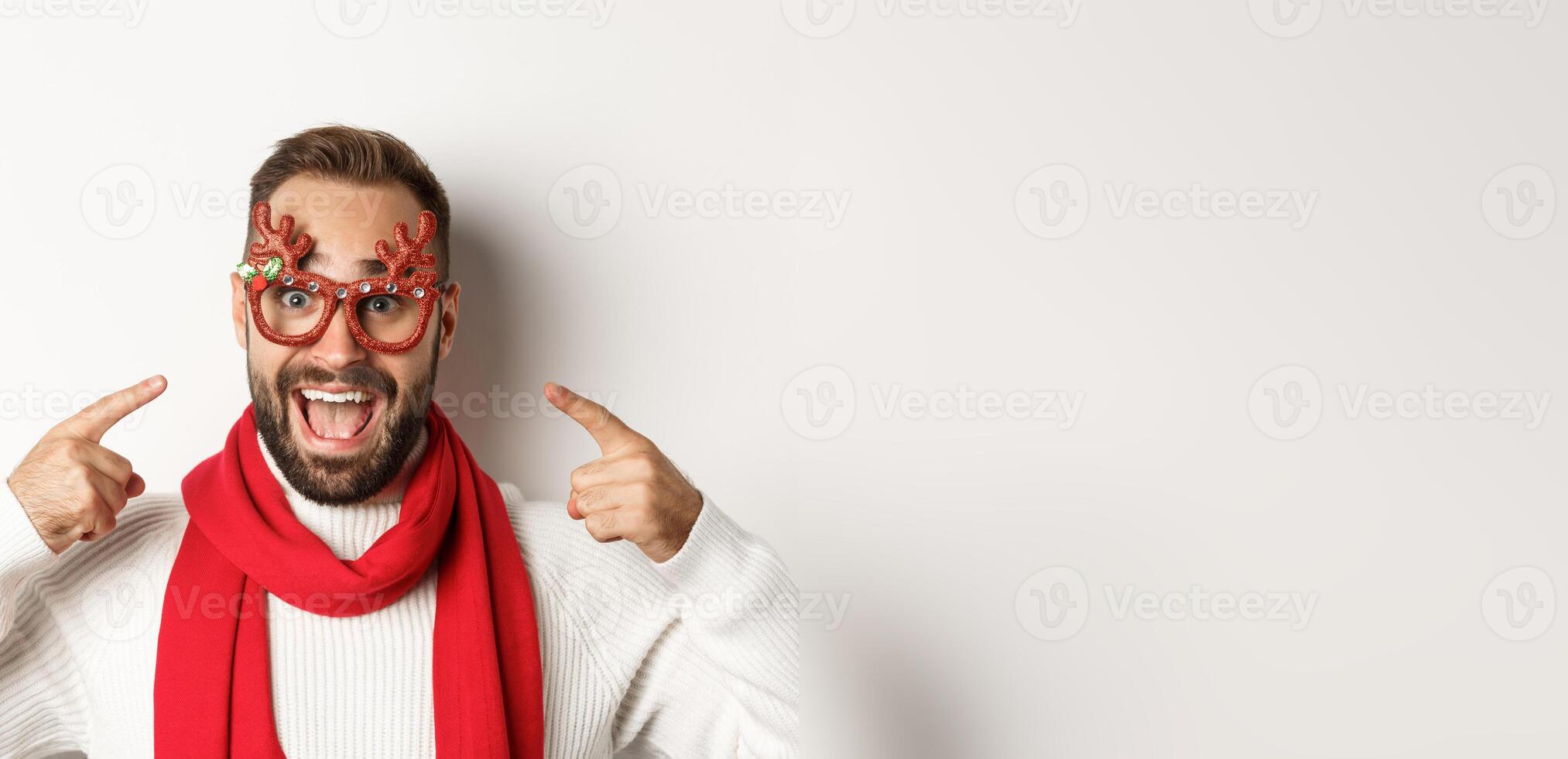 Christmas, New Year and celebration concept. Excited bearded man pointing at his party glasses, having fun and standing over white background photo