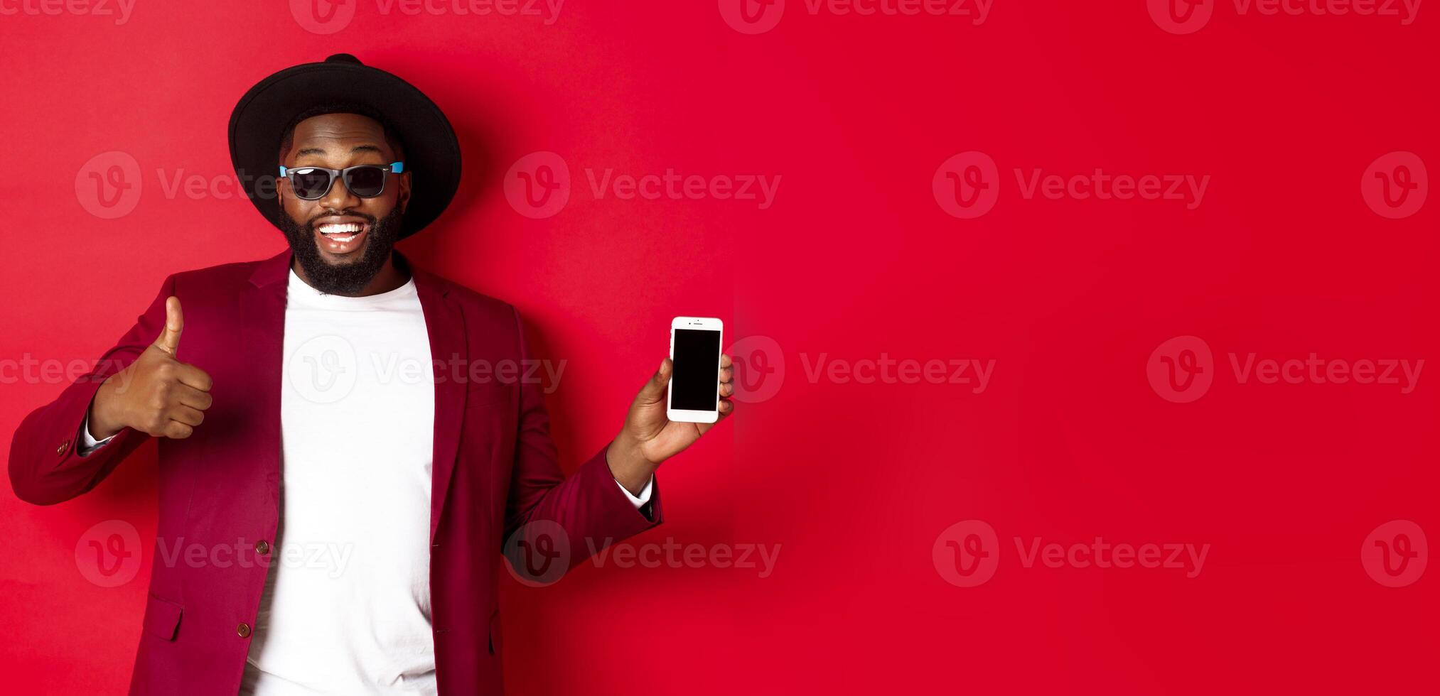 Handsome and stylish Black man showing phone screen and thumb up at camera, recommending online store app, red background photo