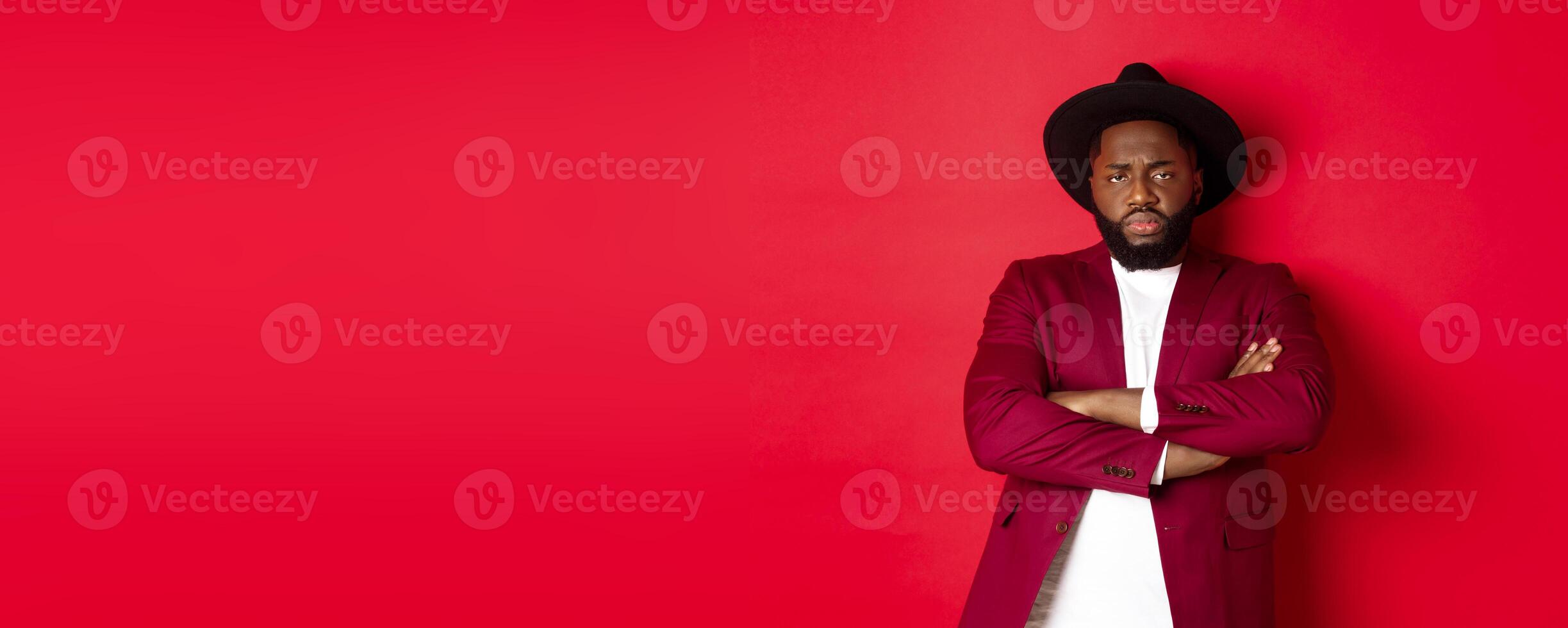 Reluctant and upset Black man waiting for apology, cross arms on chest and looking offended at camera, red background photo