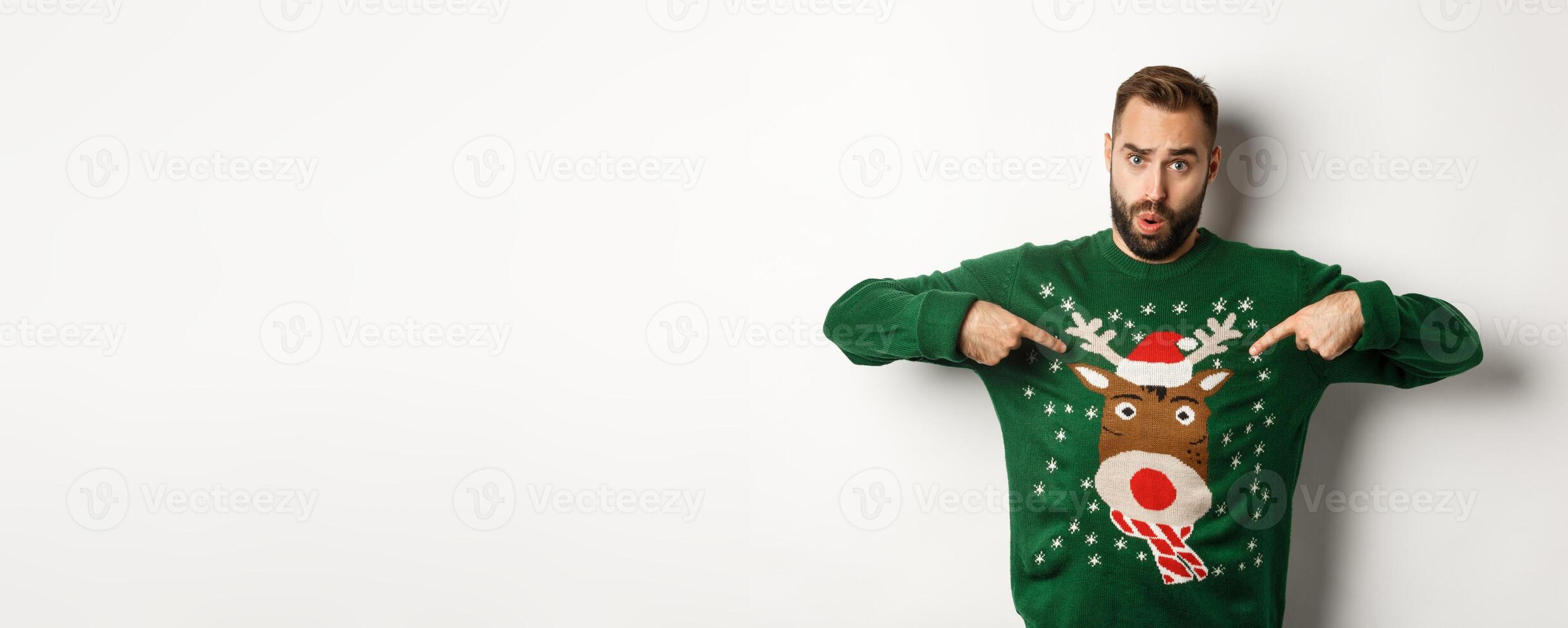 New year celebration and winter holidays concept. Man pointing at his funny christmas sweater and looking confused, standing over white background photo