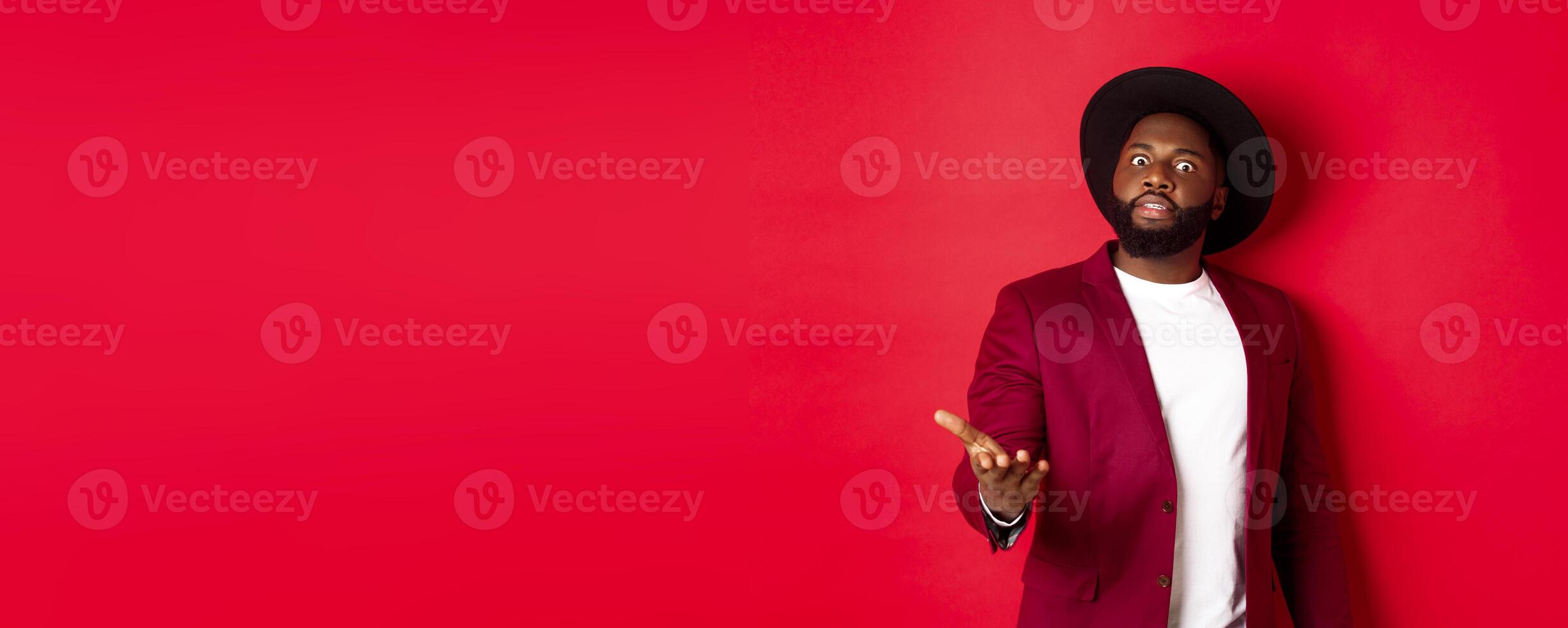 Shocked african american man staring at camera and pointing at something upsetting, standing in party jacket and hat, red background photo