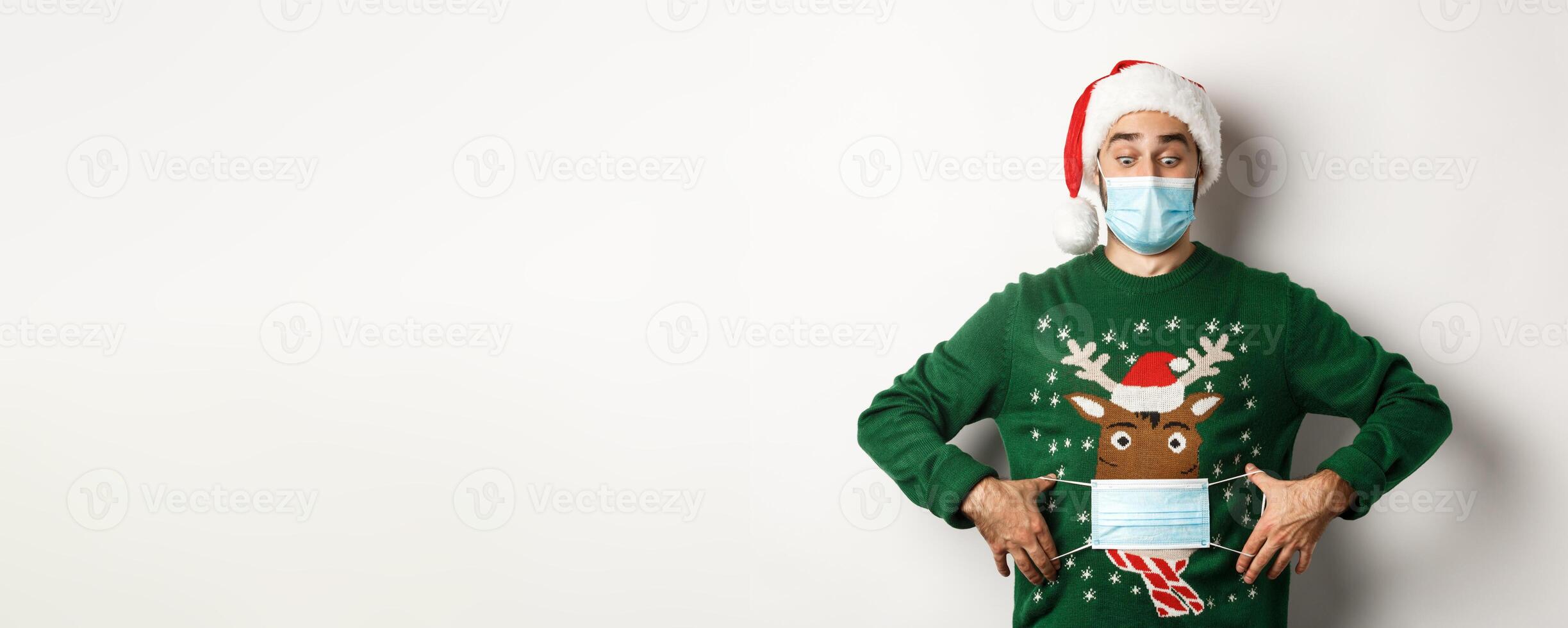 Concept of covid-19 and Christmas holidays. Funny man put on face mask on his sweater deer, standing over white background photo