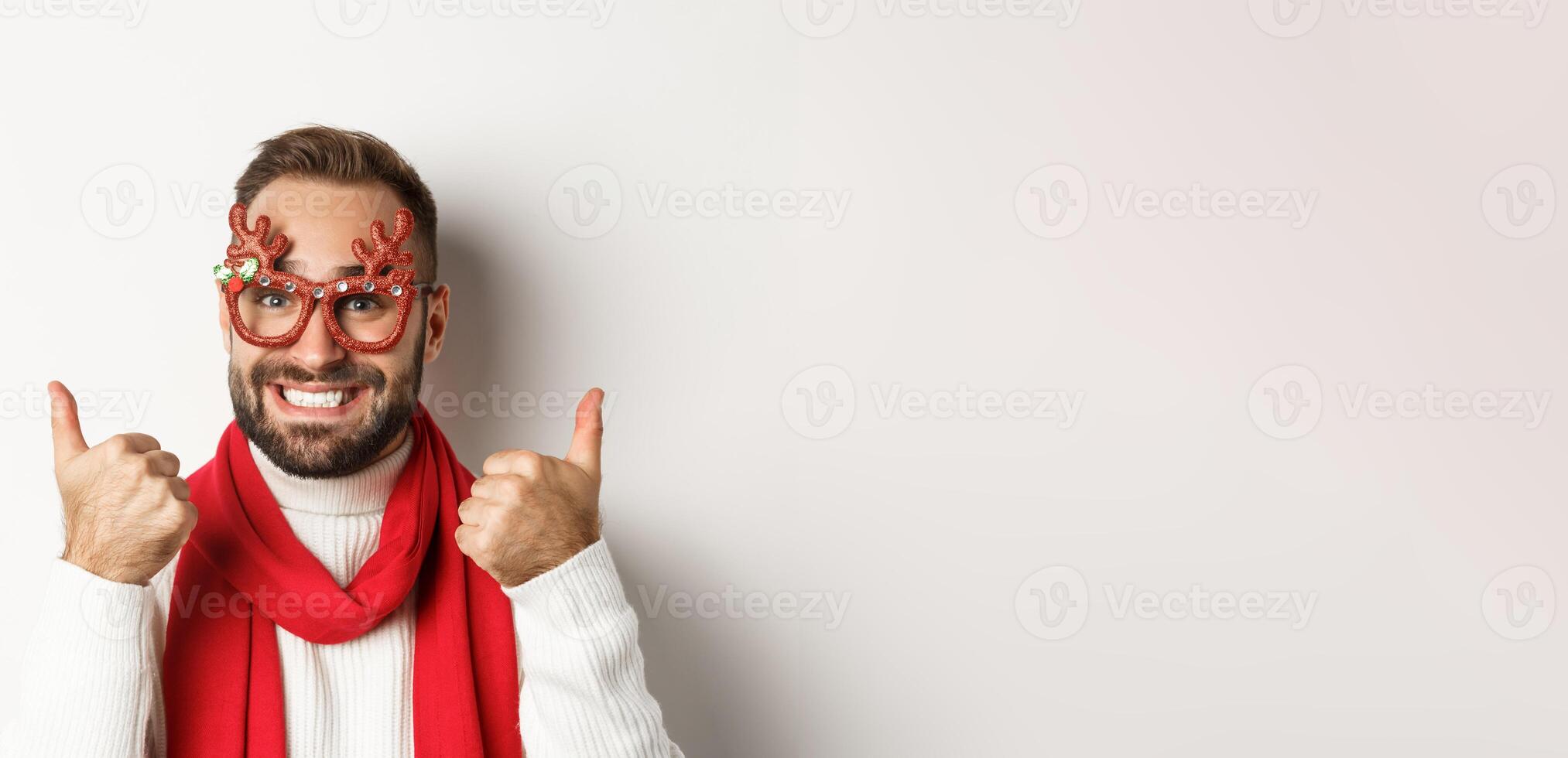 Christmas, New Year and celebration concept. Happy and satisfied man with beard, wearing party glasses, showing thumbs up in approval or like, standing over white background photo