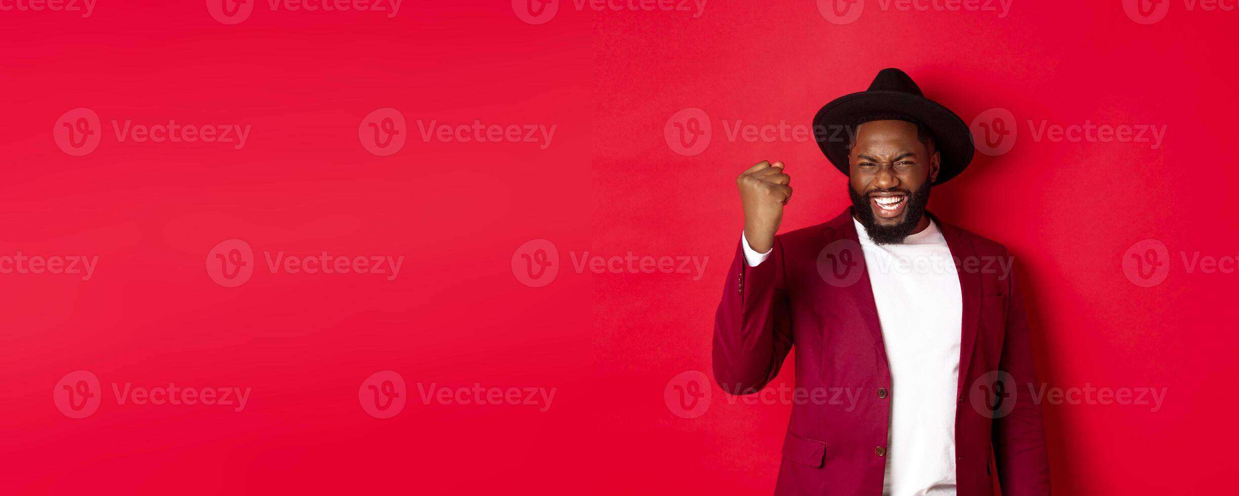 Fashion and party concept. Excited Black man rejoicing of winning, making fist pump gesture and say yes with satisfaction, standing against red background photo