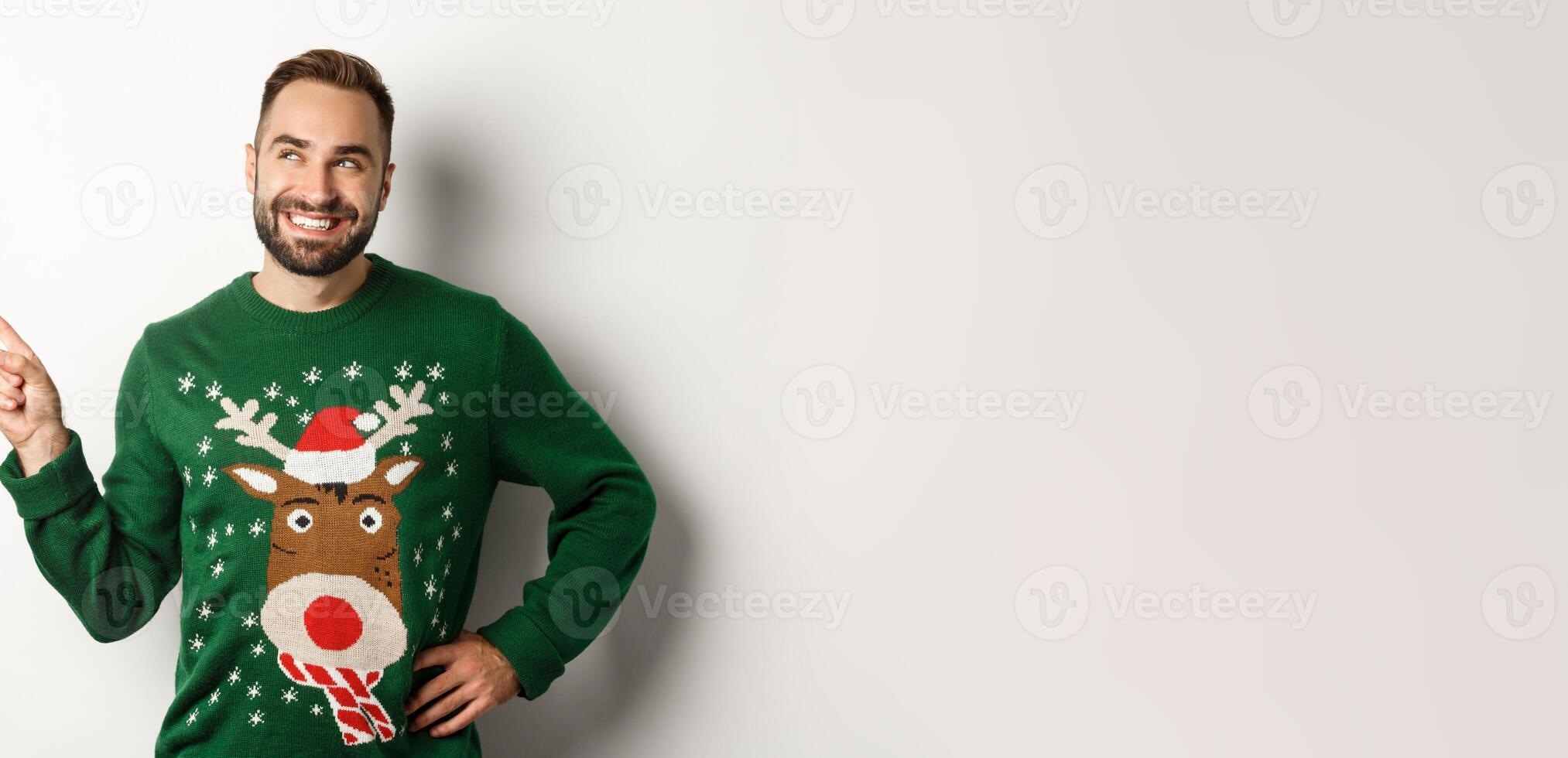 New year celebration and winter holidays concept. Handsome smiling man in green christmas sweater, pointing and looking at upper left corner logo store, white background photo