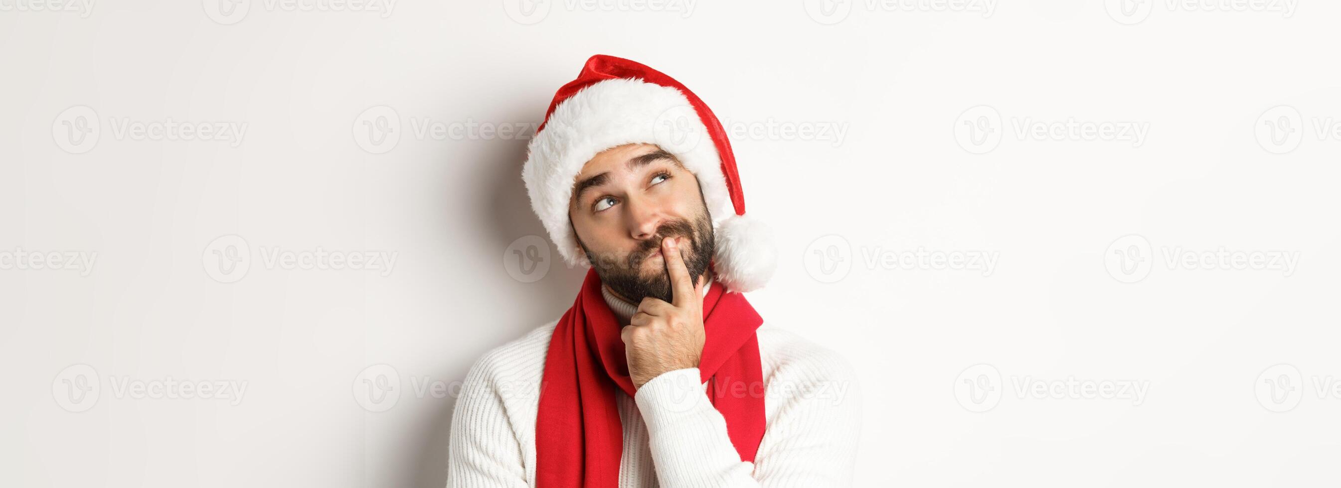 New Year party and winter holidays concept. Close-up of bearded man in Santa hat looking thoughtful at upper left corner, thinking or making decision, choosing christmas gift, white background photo