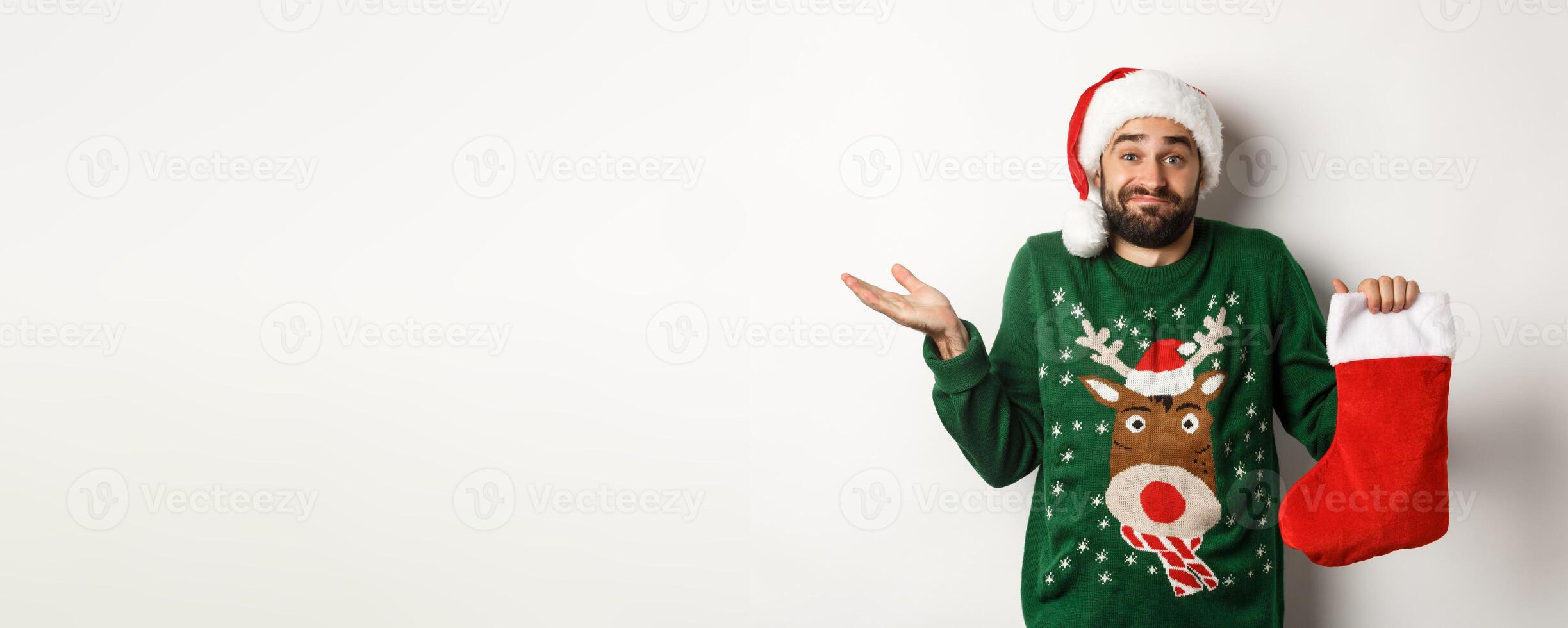 Xmas and winter holidays concept. Confused guy in Santa hat holding Christmas sock and shrugging, standing clueless against white background photo