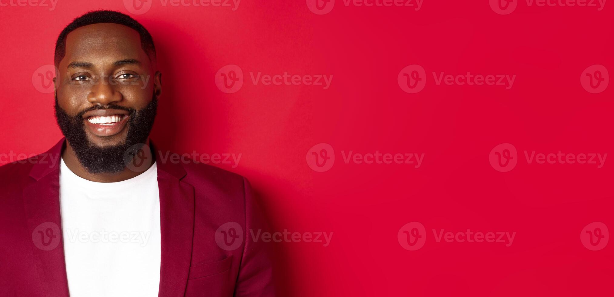 Close-up of handsome and stylish Black man wearing party blazer, smiling happy at camera, standing against red background photo