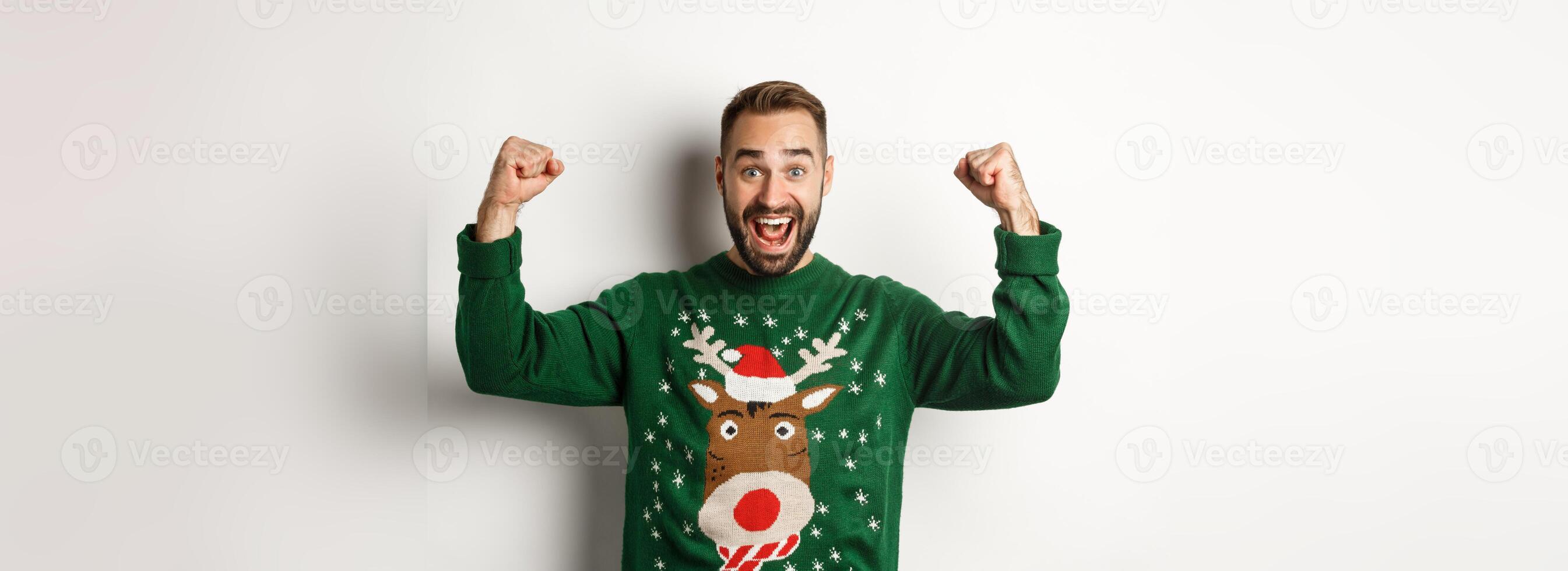 New Year, holidays and celebration. Relieved and excited man in Christmas sweater, raising hands up and celebrating, triumphing, standing over white background photo
