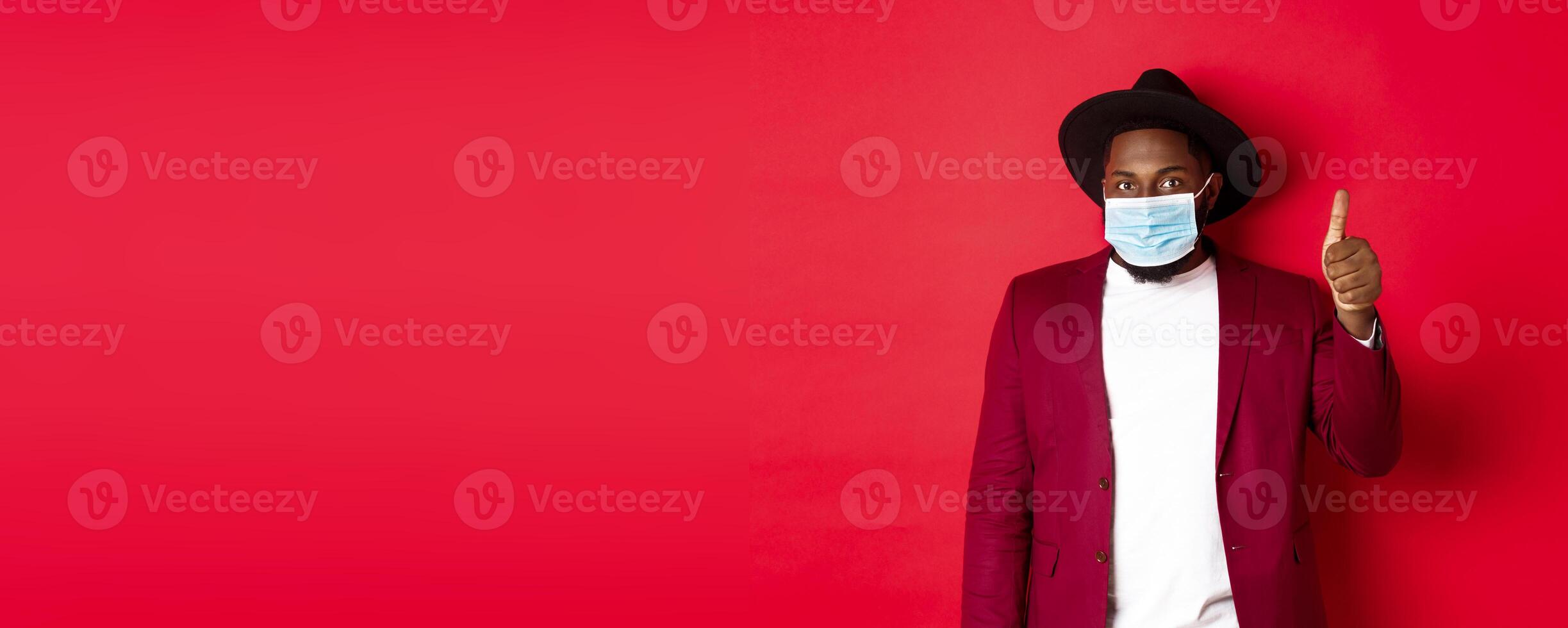 Covid-19 and fashion concept. Stylish african american man in hat and blazer, wearing face mask and showing thumb up, standing over red background photo