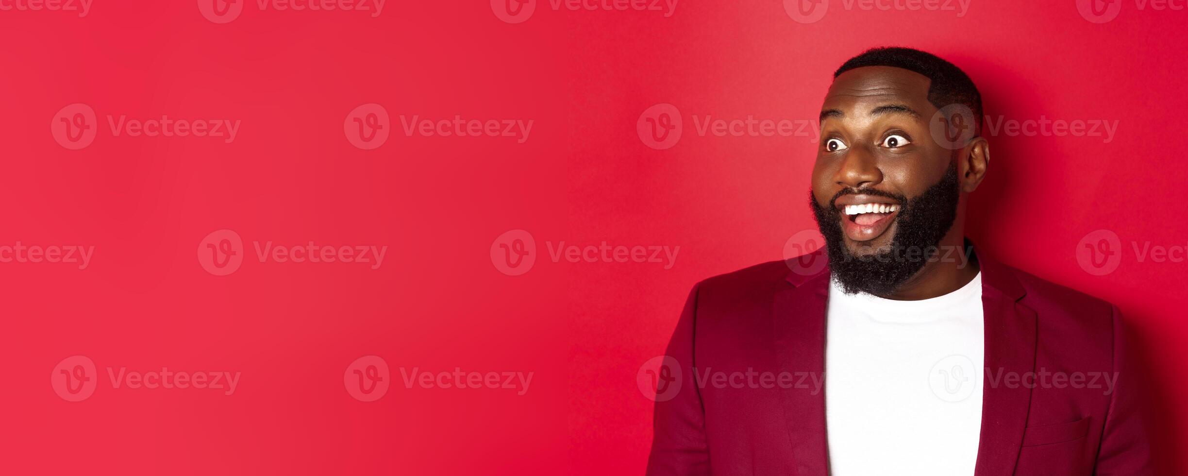 Close-up of excited african american guy looking left, smiling with amazement and joy, standing in blazer against red background photo