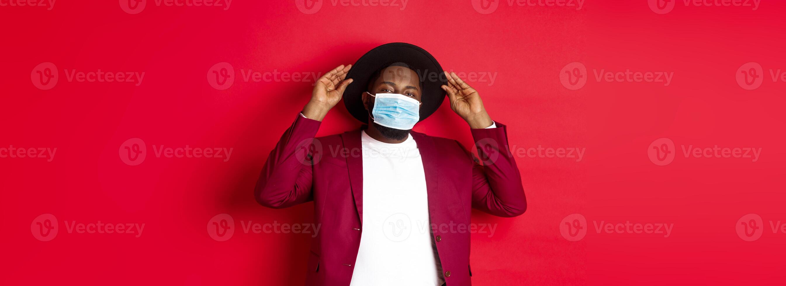 Covid-19, quarantine and holidays concept. Handsome and stylish african american man in face mask, put hat on head and looking sassy, dress-up for party, standing over red background photo