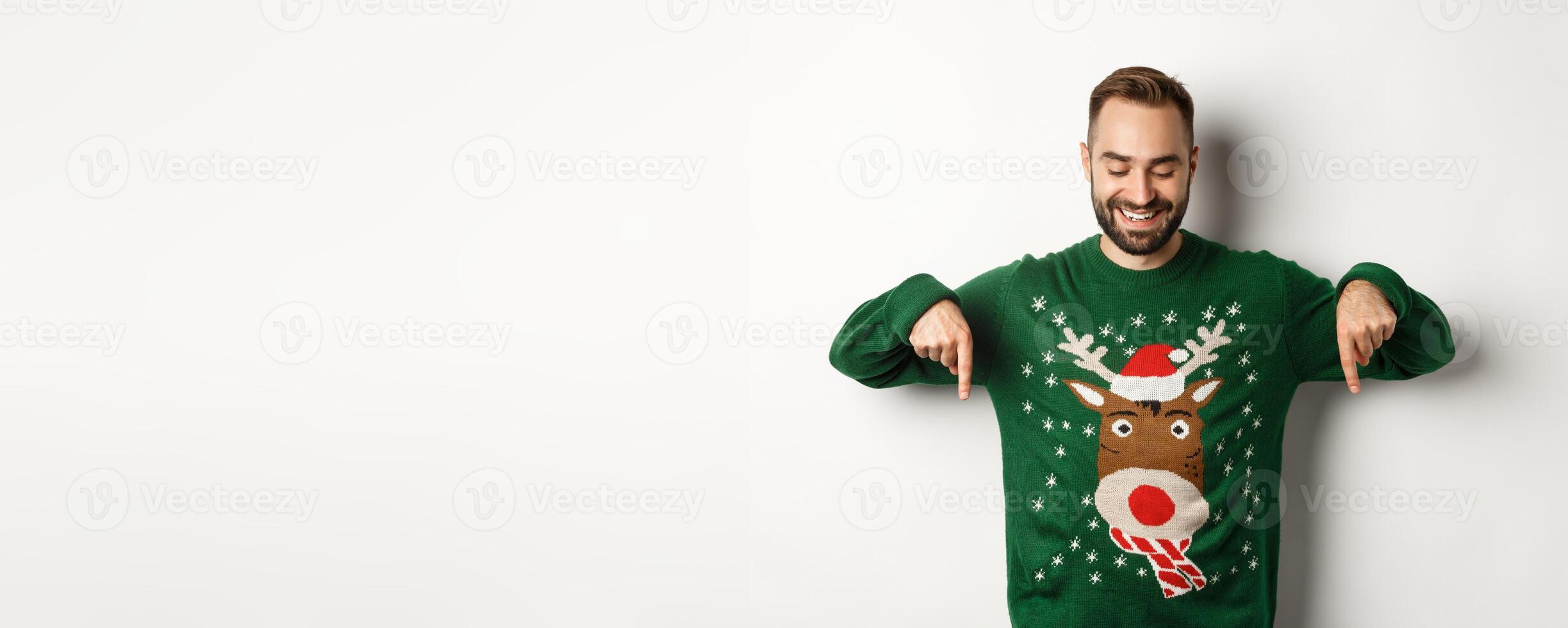 Winter holidays and christmas. Happy caucasian man in green sweater, pointing fingers down and looking satisfied, standing over white background photo