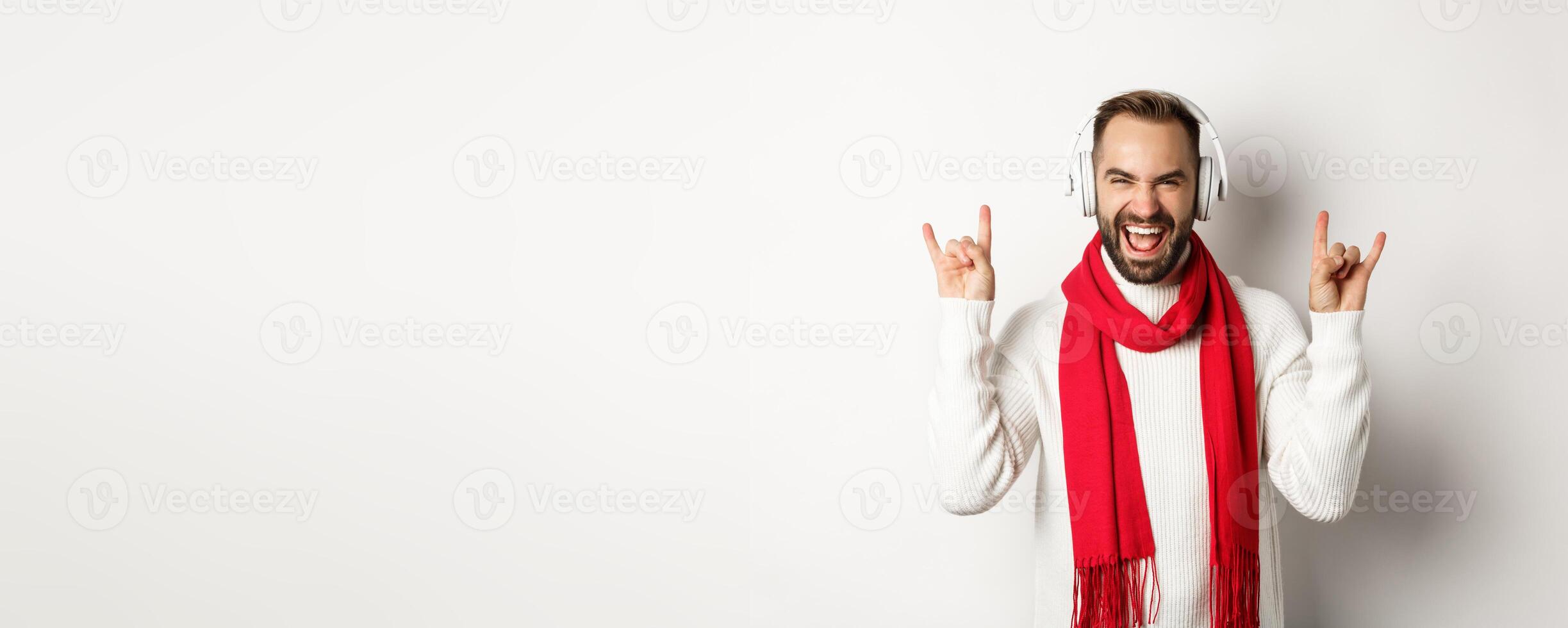 Joyful guy listening heavy metal in headphones, showing rock-on gesture and shouting for joy, standing over white background photo