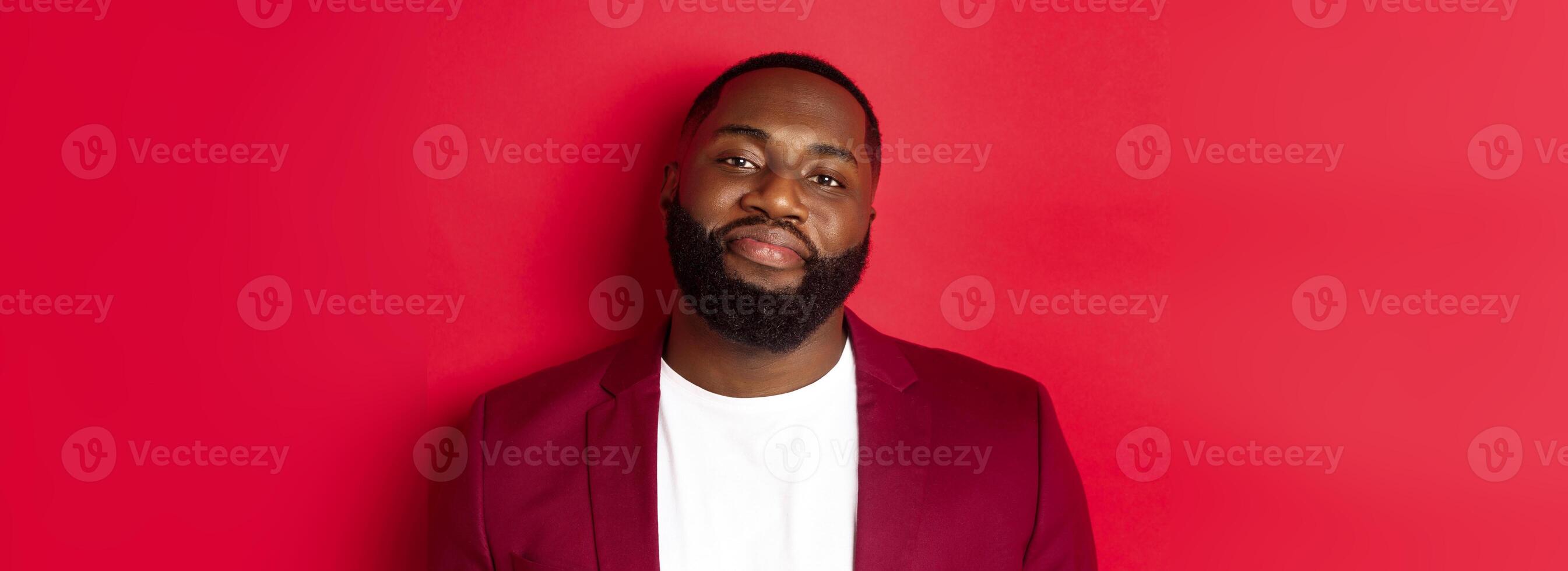 Close-up of handsome bearded plump guy smiling at camera shy, standing in stylish blazer on t-shirt, standing over red background photo