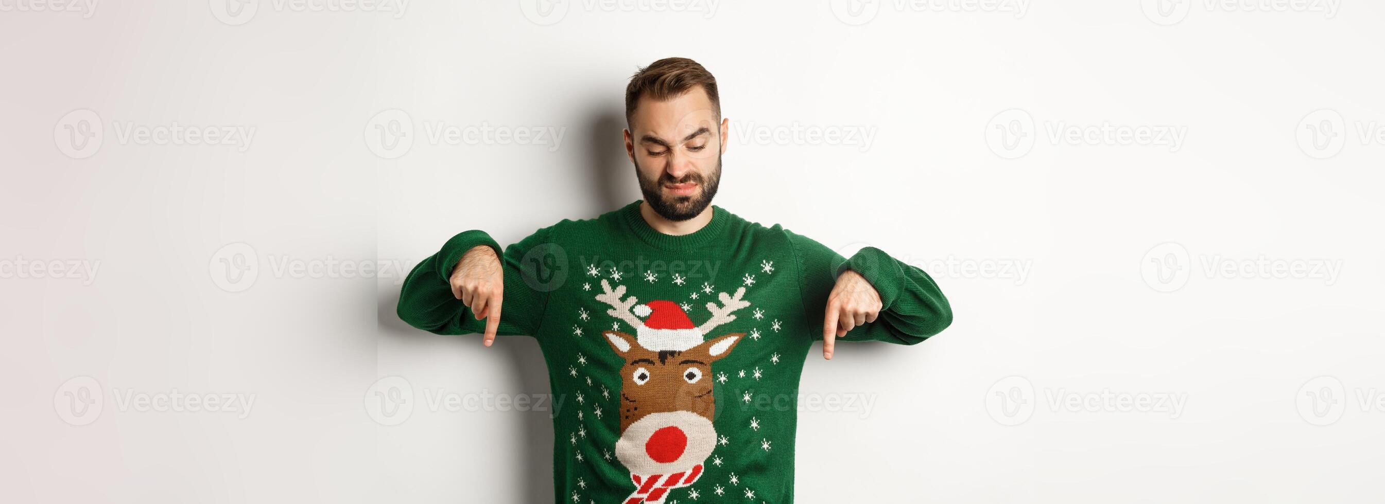 Winter holidays and christmas. Skeptical bearded man in green sweater, pointing and looking down displeased, standing over white background photo