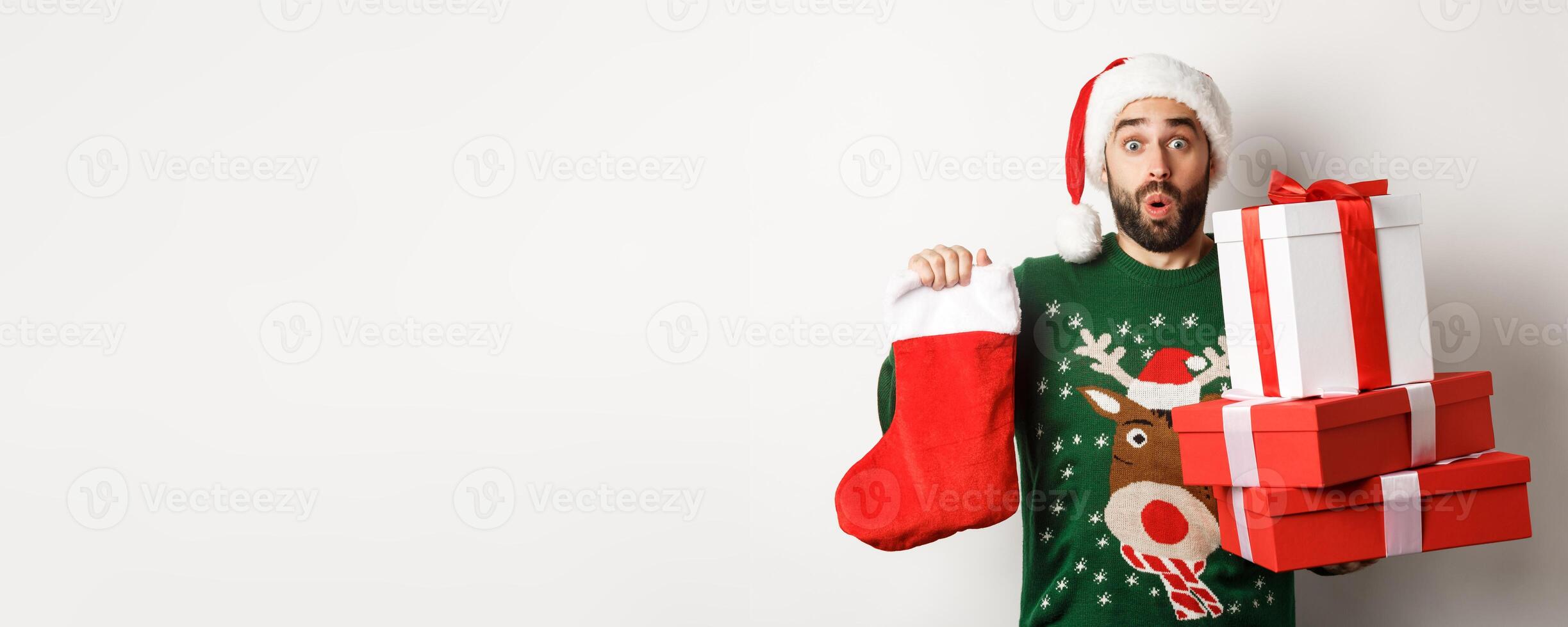 Xmas and winter holidays concept. Excited man holding christmas sock and gift boxes, celebrating New Year, bringing presents under tree, standing over white background photo