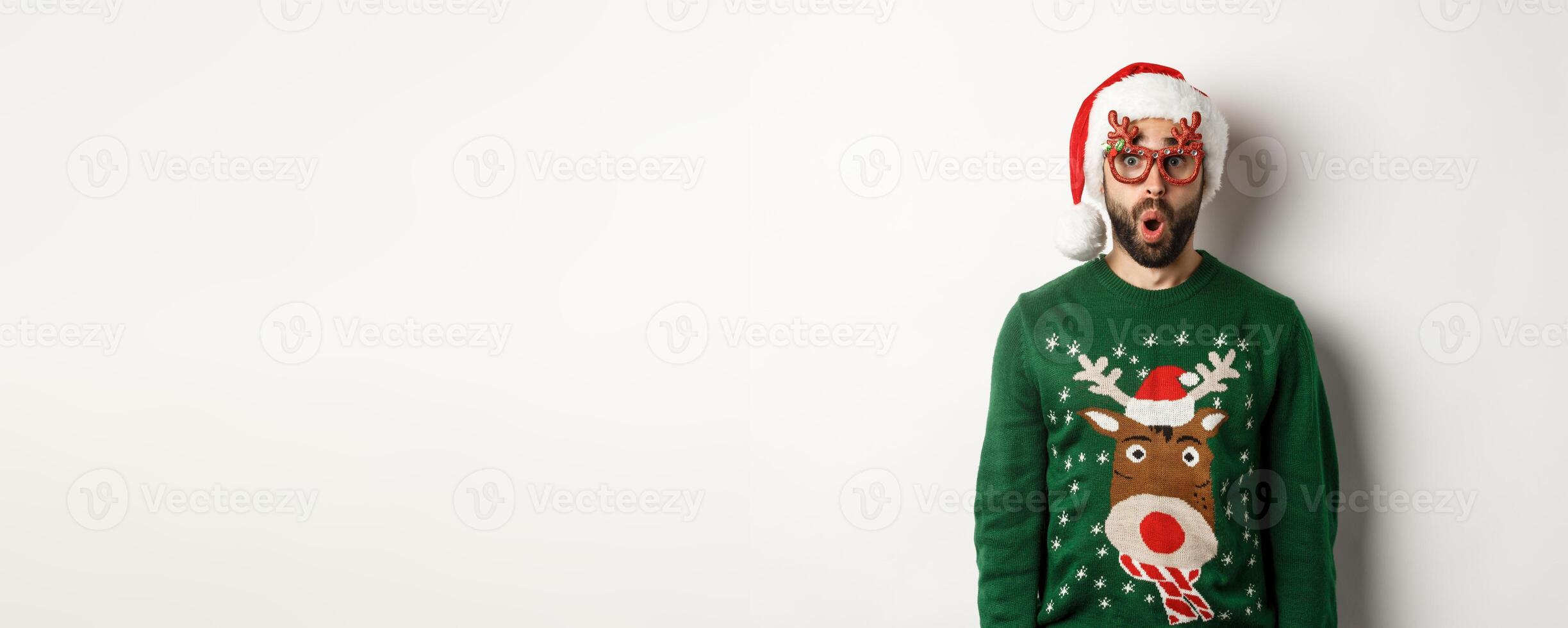 Christmas holidays, celebration concept. Surprised man in Santa hat and party glasses looking at something awesome, standing over white background photo
