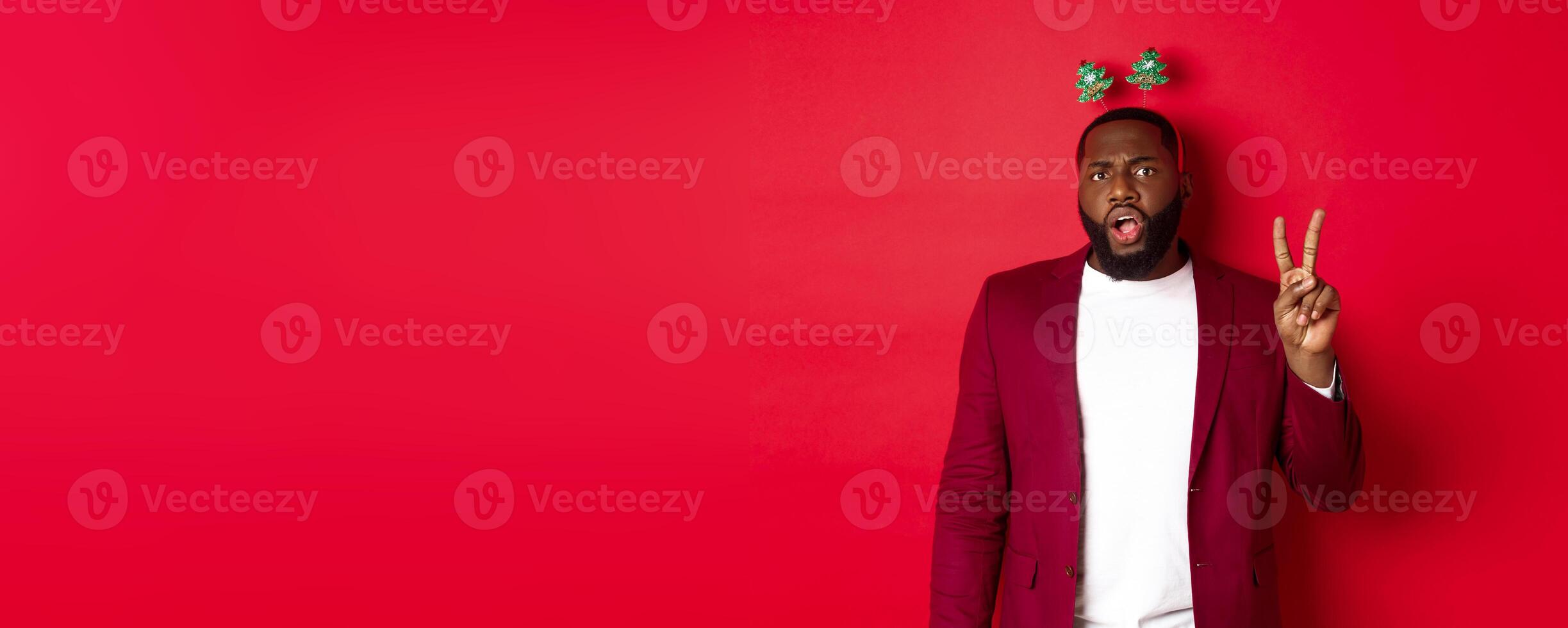 Merry Christmas. Masculine black man in costume and silly party headband, showing peace sign but looking serious, standing over red background photo