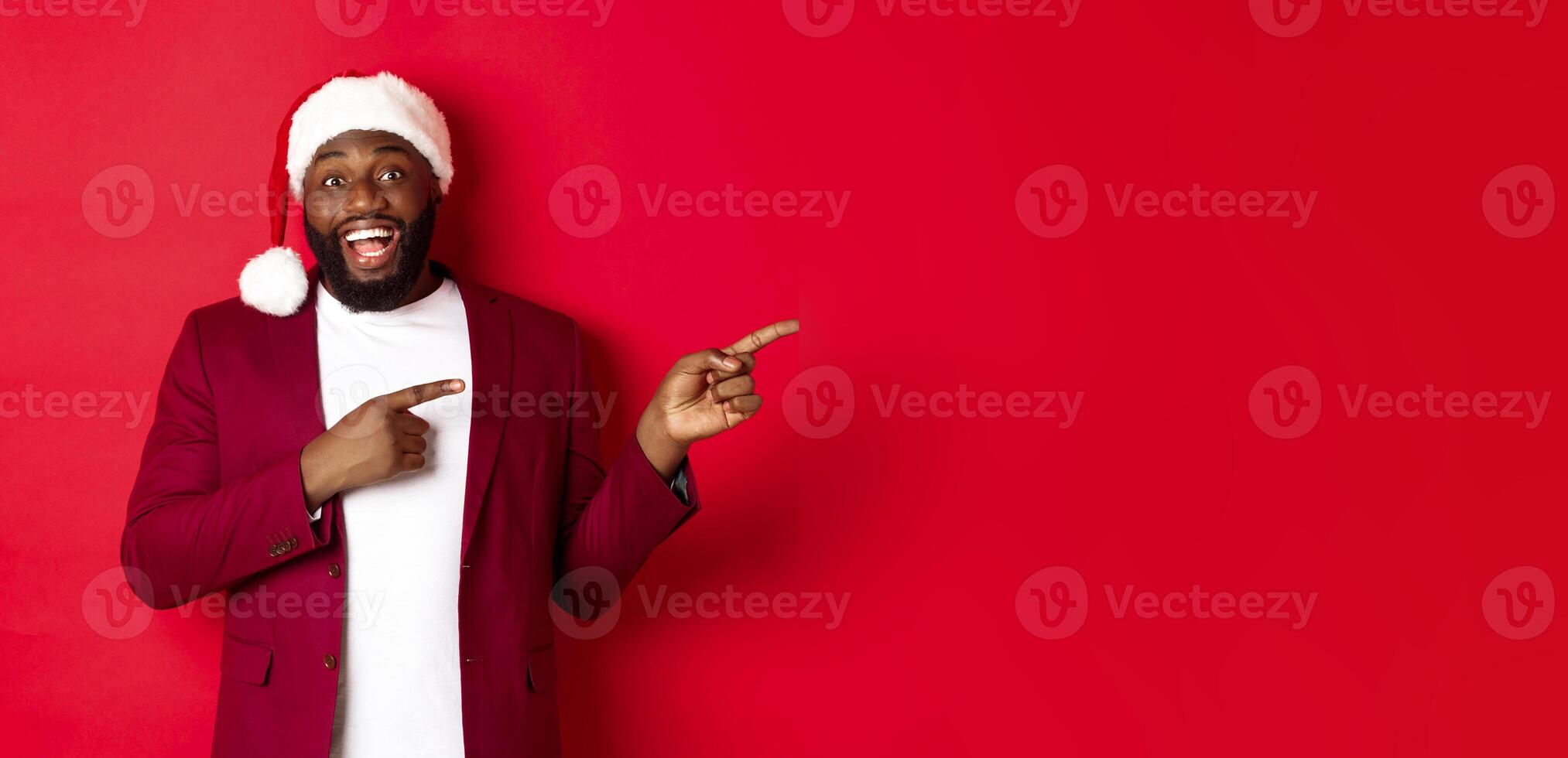 Christmas, party and holidays concept. Cheerful Black man smiling, pointing fingers right and showing advertisement, standing over red background photo