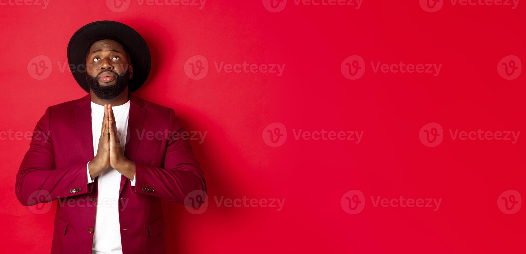 Hopeful and sad Black man talking to God, holding hands in pray and looking up, pleading, begging for help, standing against red background photo