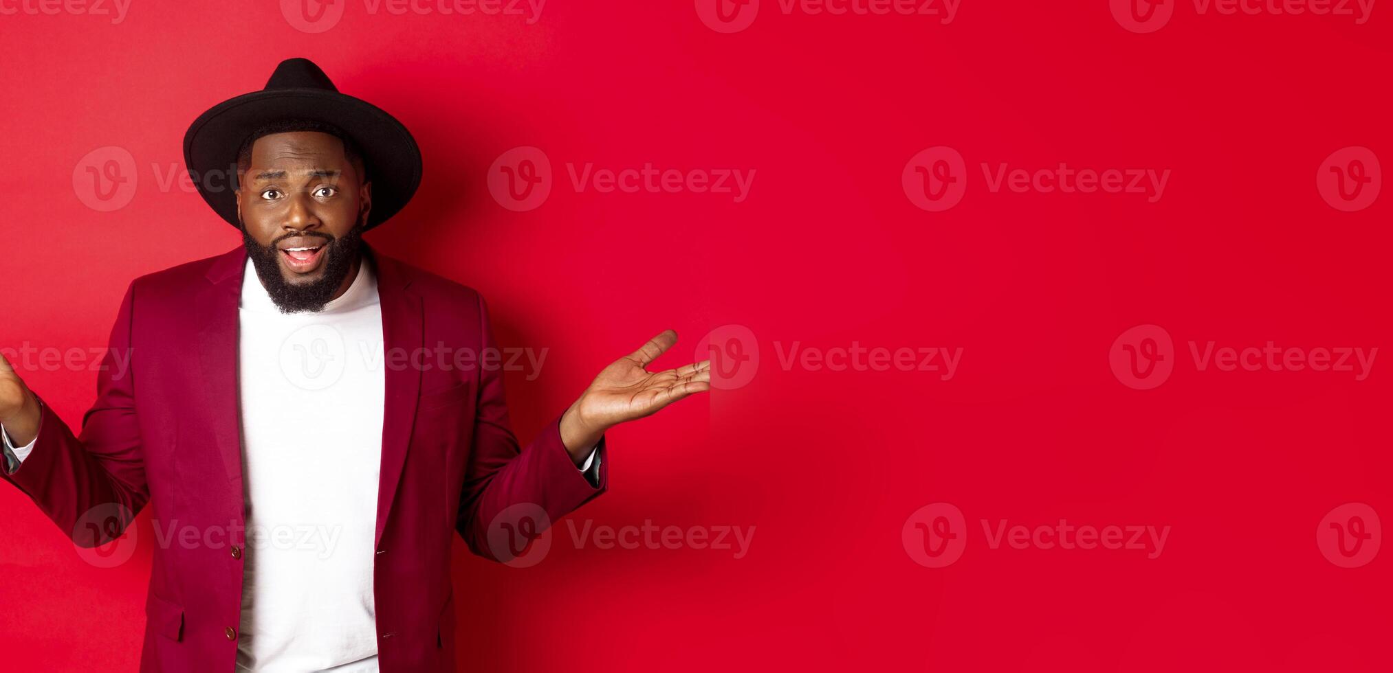 Image of confused Black man asking question, spread hands sideways and staring camera clueless, standing against red background photo
