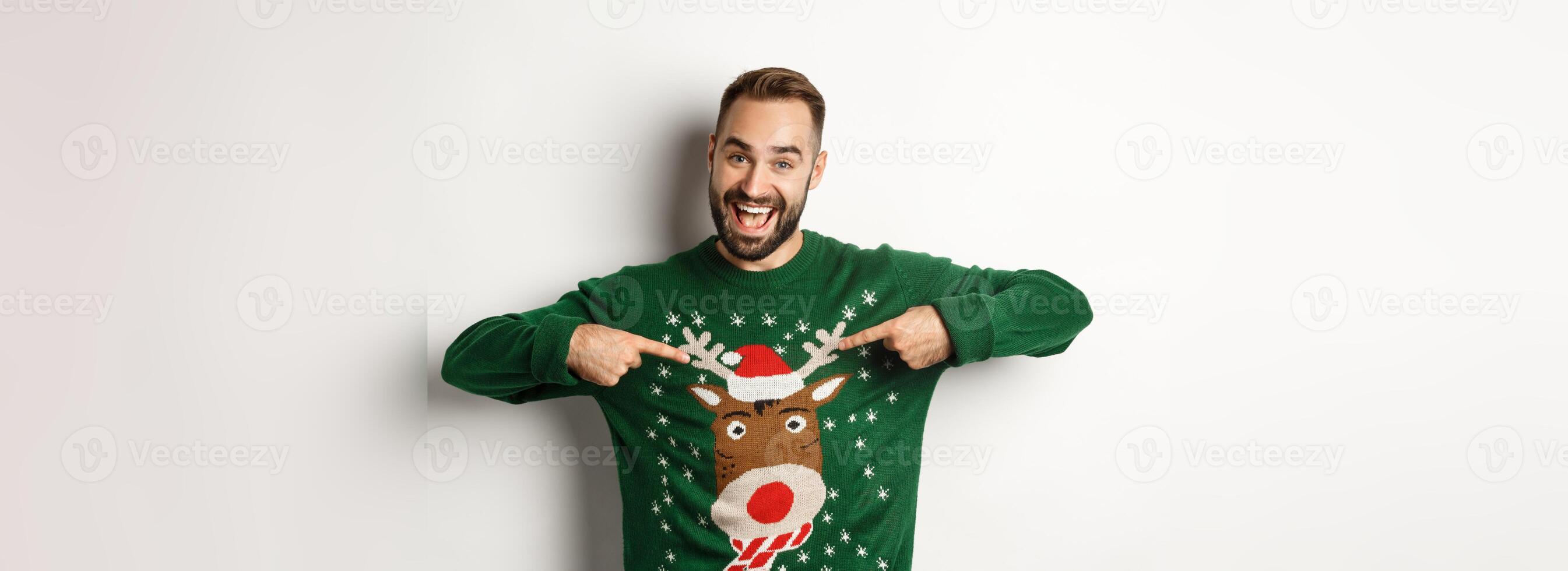 New year celebration and winter holidays concept. Cheerful man pointing at his funny Christmas sweater and smiling, white background photo