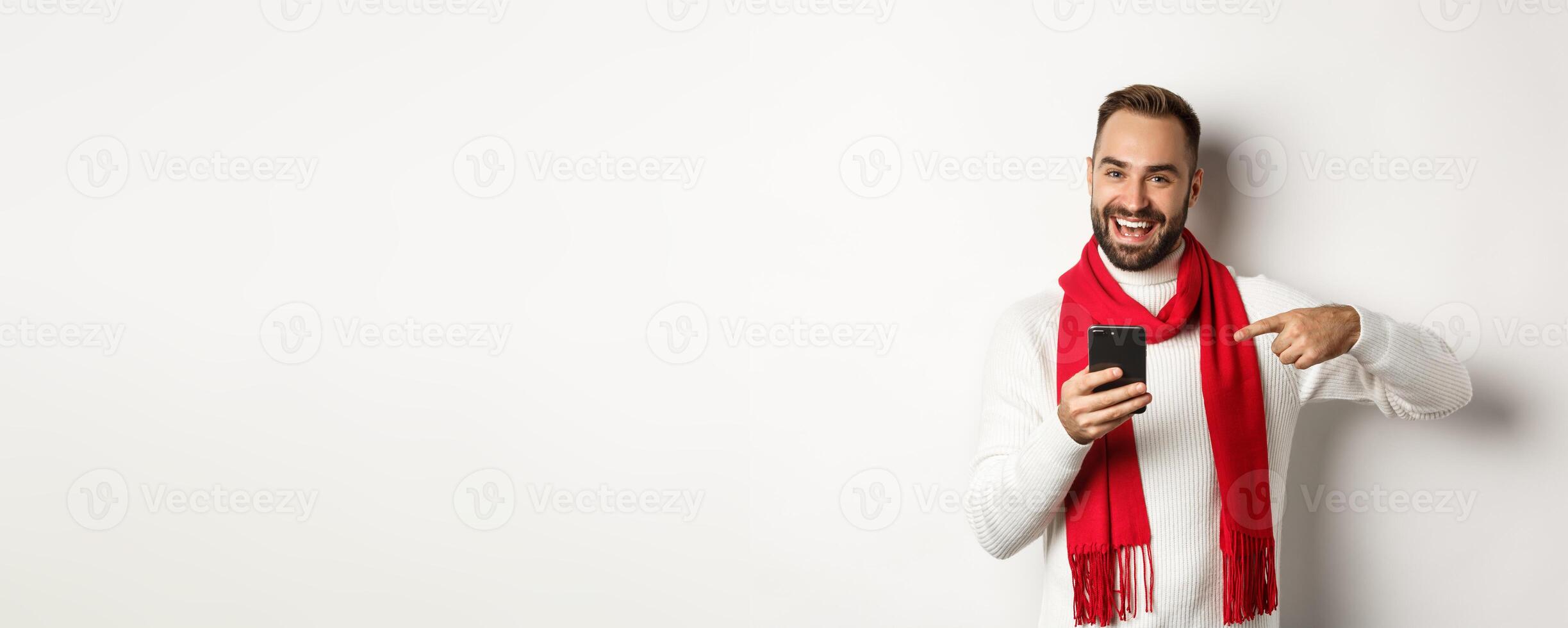 Satisfied bearded guy pointing finger at smartphone, checking out internet promo offer on mobile phone, standing over white background photo
