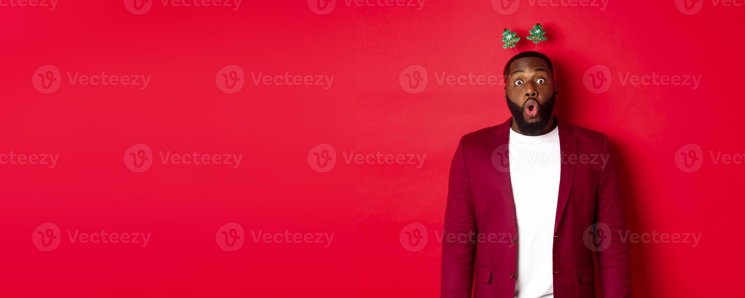 Merry Christmas. Happy african american man celebarting New Year, wearing party headband and making funny faces , standing over red background photo