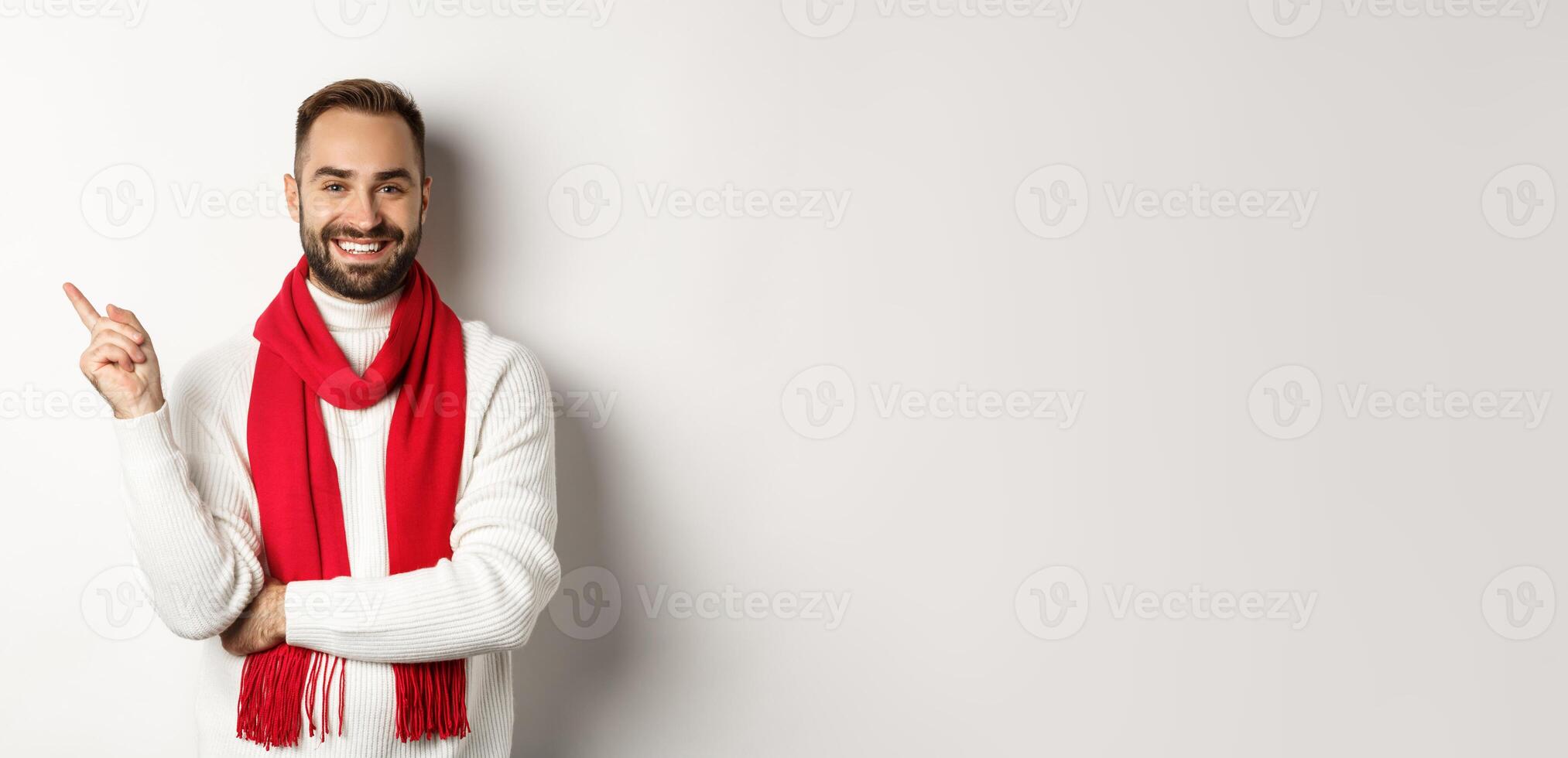 Christmas shopping and winter holidays concept. Young man with beard pointing finger left, showing new year advertisement, standing over white background in red scarf and sweater photo