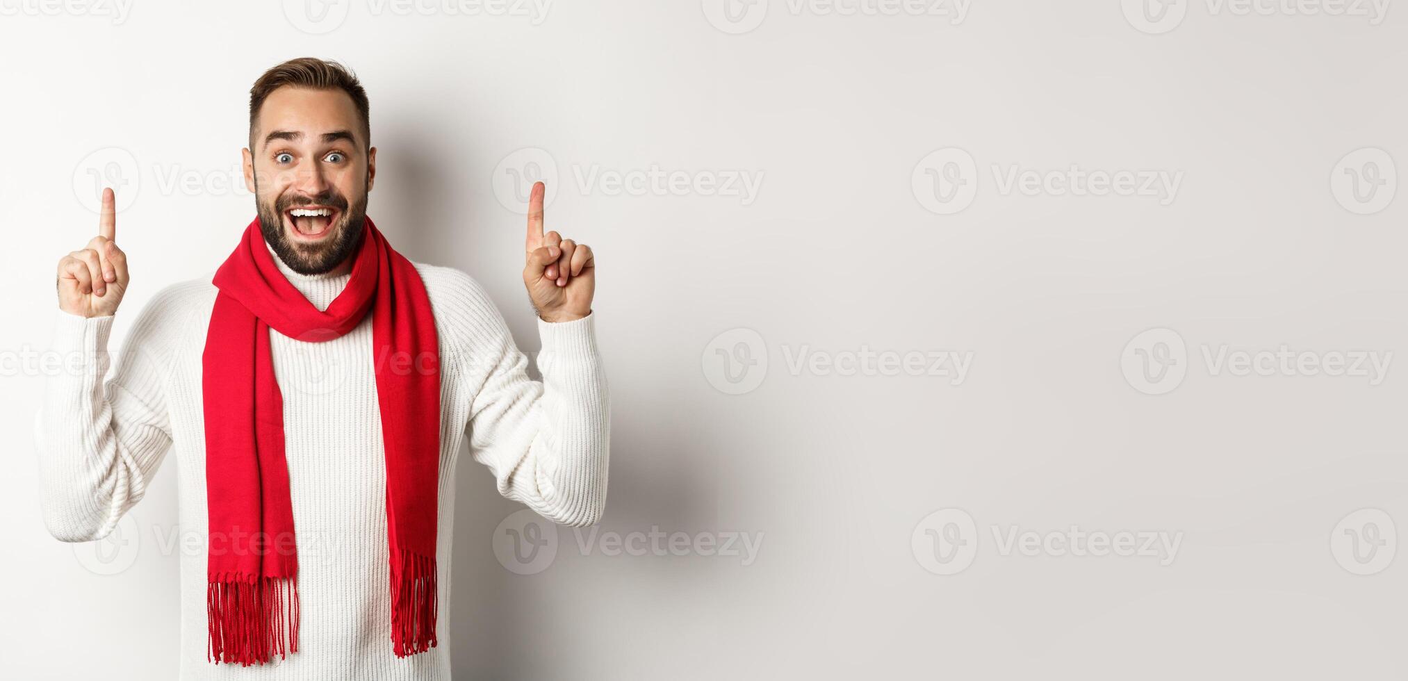 Christmas shopping and winter holidays concept. Cheerful handsome man showing promo banner, pointing fingers up at copy space, wearing red scarf and sweater, white background photo