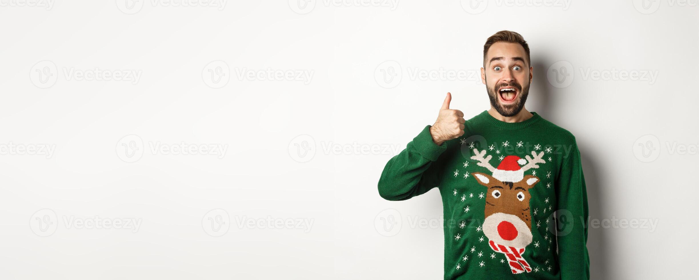 Winter holidays and christmas. Excited caucasian guy showing thumb up and looking amazed, standing in green sweater against white background photo