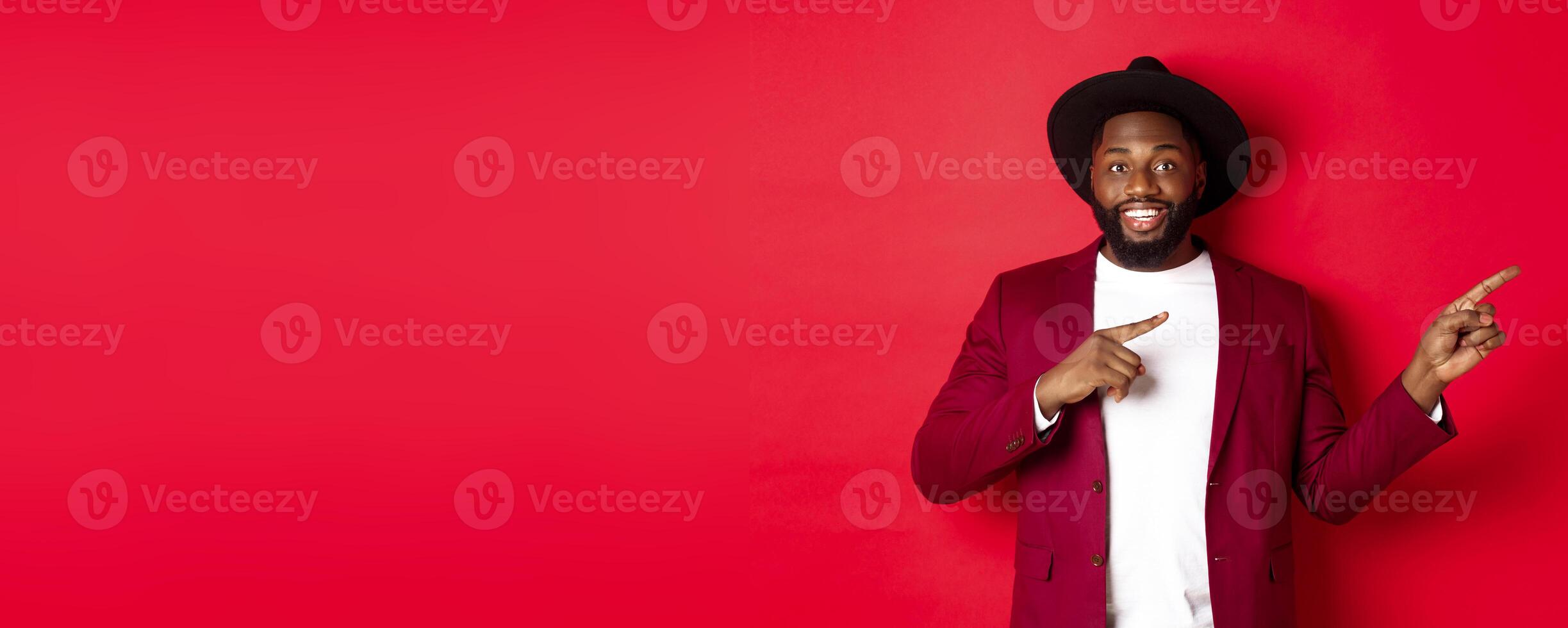 Winter holidays and shopping concept. Stylish african american man pointing fingers right at copy space for logo, standing over red background photo