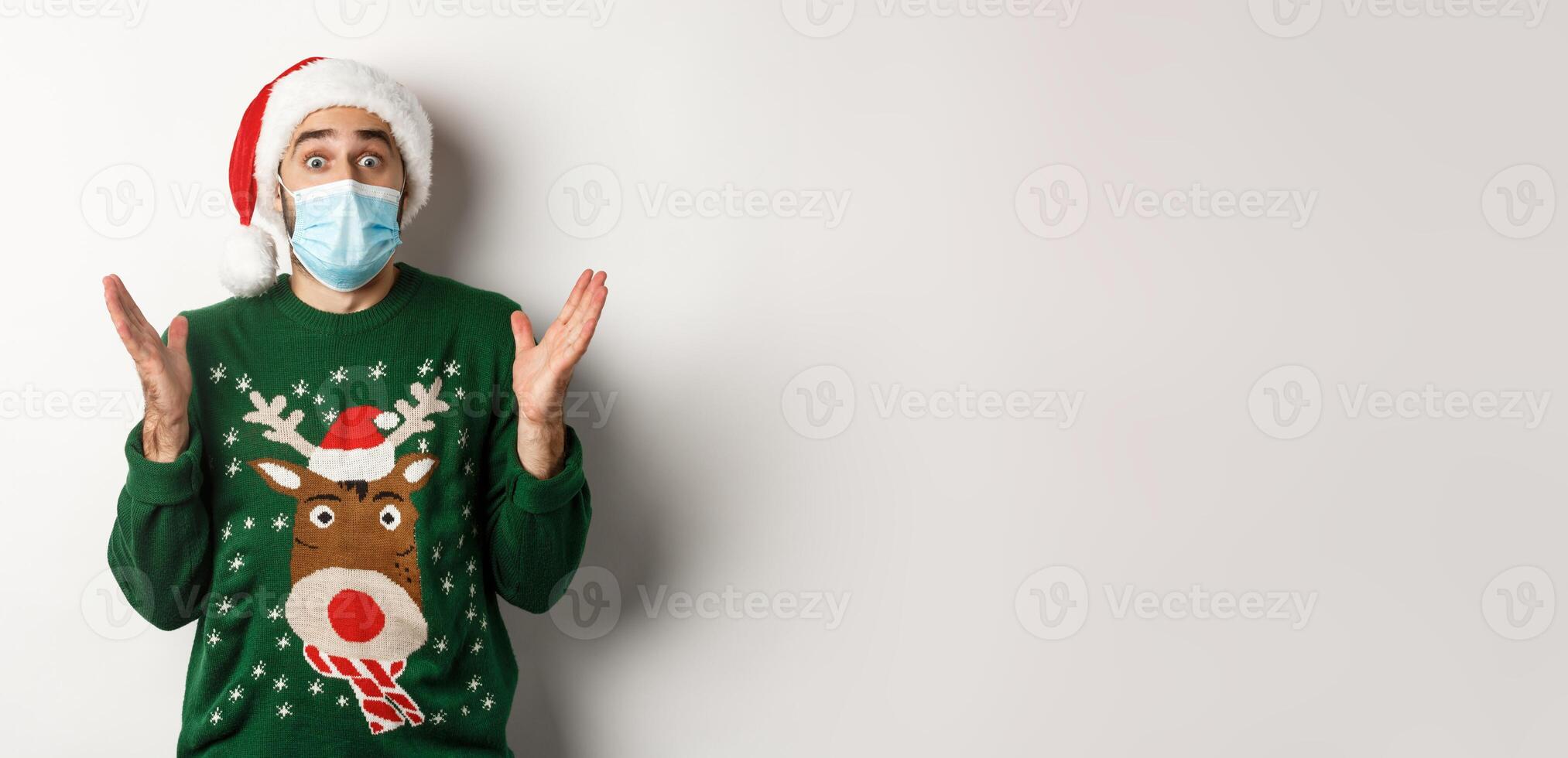 Christmas during pandemic, covid-19 concept. Surprised guy in medical mask, Santa hat and sweater celebrating New Year party, standing over white background photo