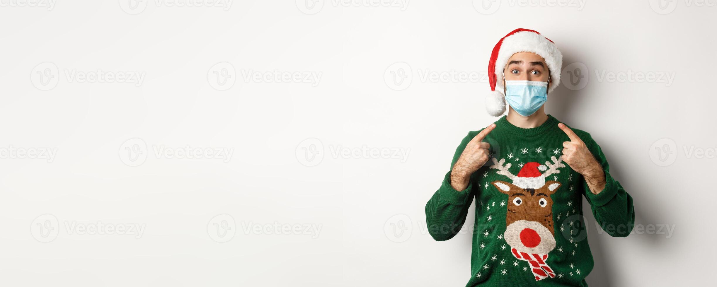 Christmas during pandemic, covid-19 concept. Man in in Santa hat pointing at his face mask, celebrating New Year and social distancing, white background photo