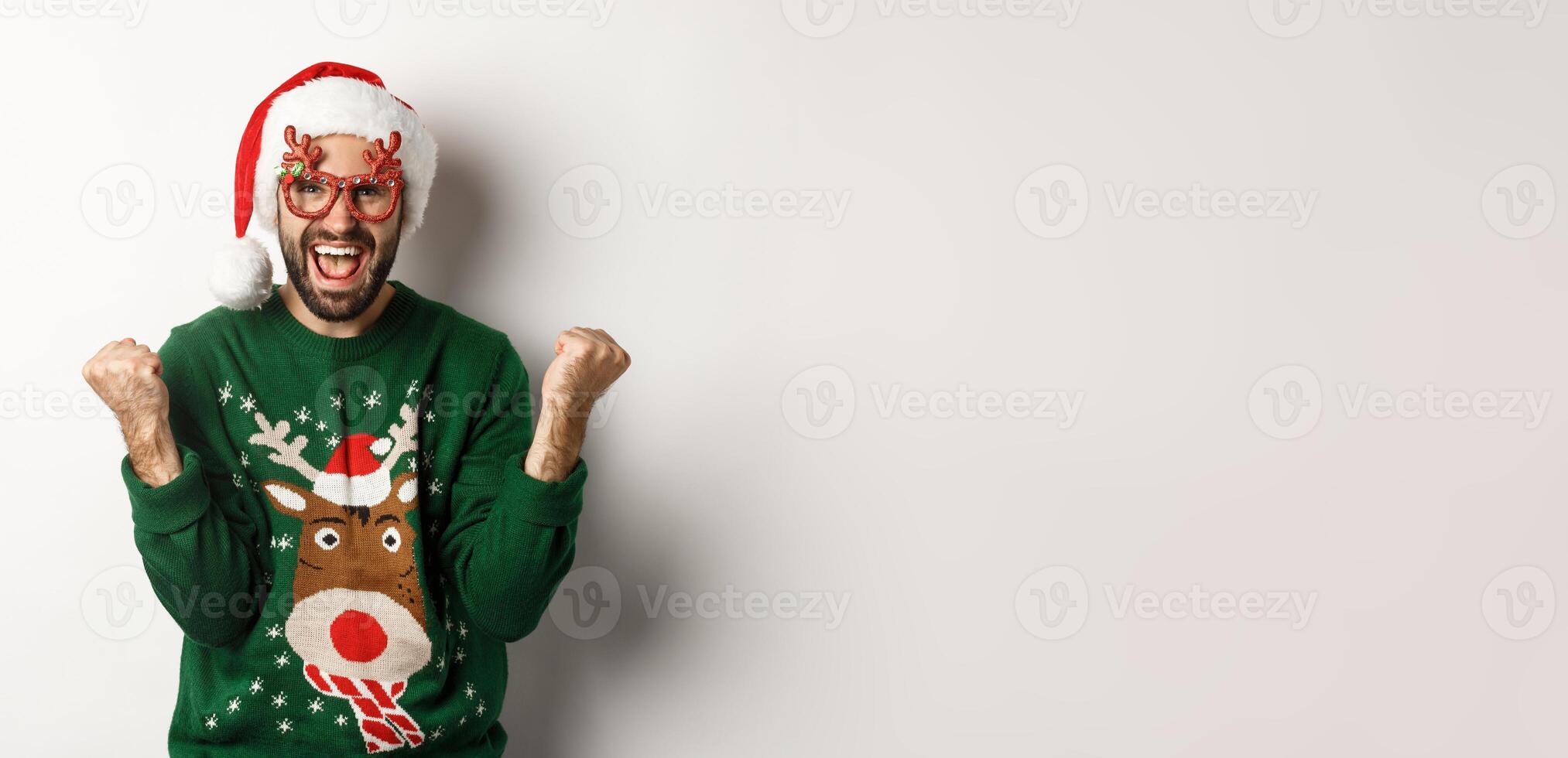 Christmas holidays, celebration concept. Happy man in Santa hat triumphing, wearing funny party glasses and rejoicing, standing over white background photo