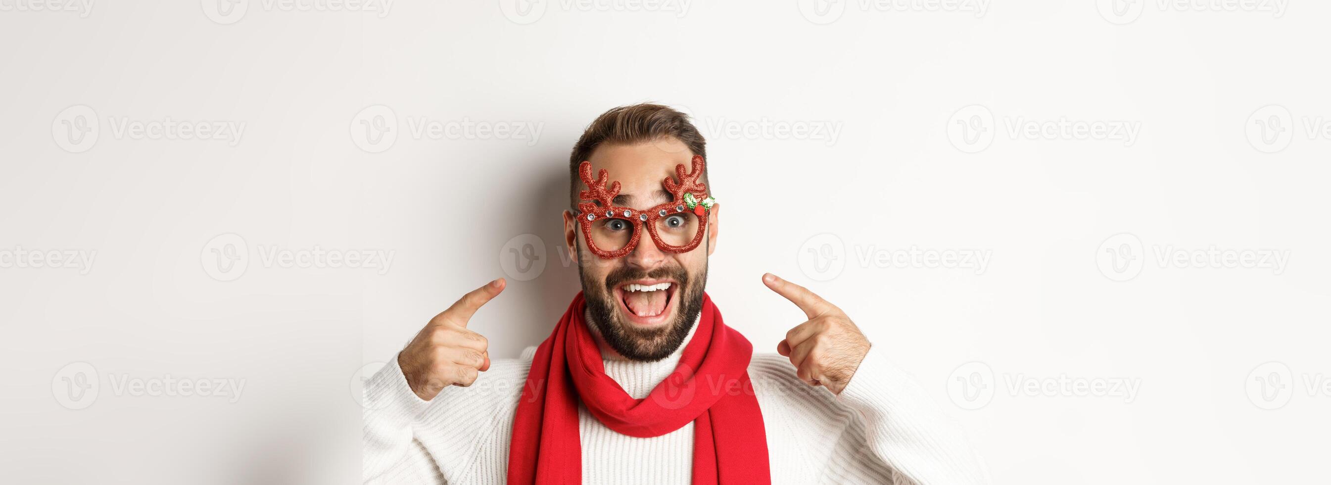 Christmas, New Year and celebration concept. Excited bearded man pointing at his party glasses, having fun and standing over white background photo