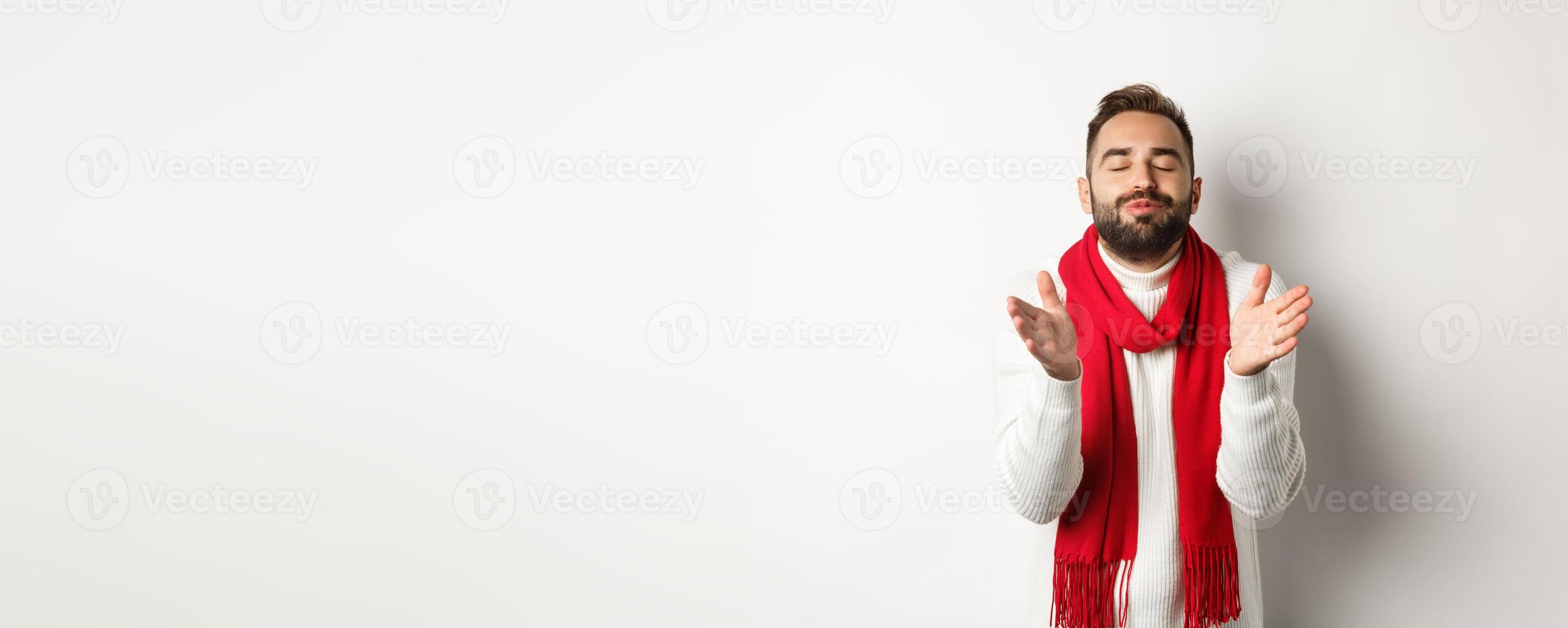 Handsome man in christmas sweater standing with closed eyes, puckered lips, leaning for kiss, standing over white background photo