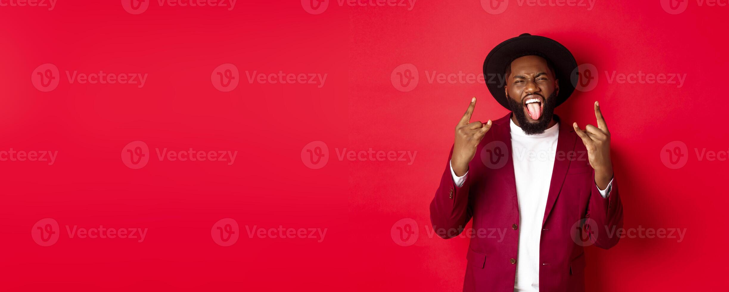 Happy african american man having fun on party, showing tongue and rock-n-roll gesture, celebrating New Year, standing against red background photo