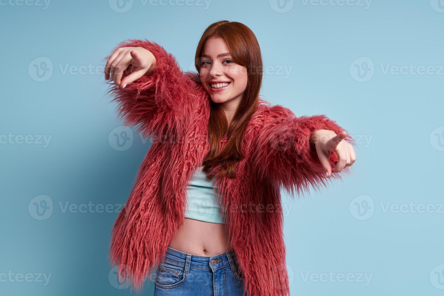 Beautiful young woman in fluffy coat pointing camera and smiling against blue background photo