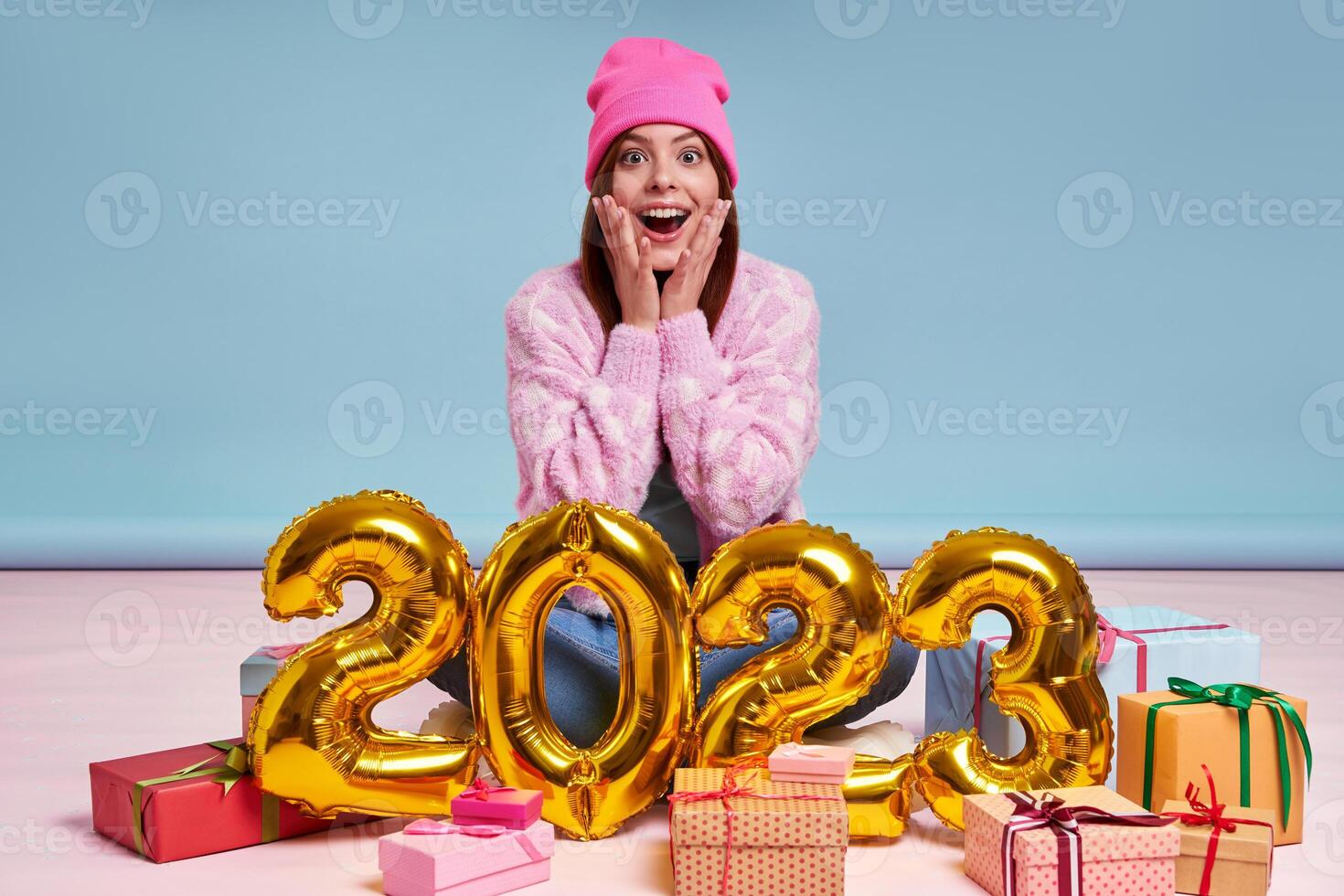 Surprised young woman sitting near the gift boxes and gold colored numbers against blue background photo