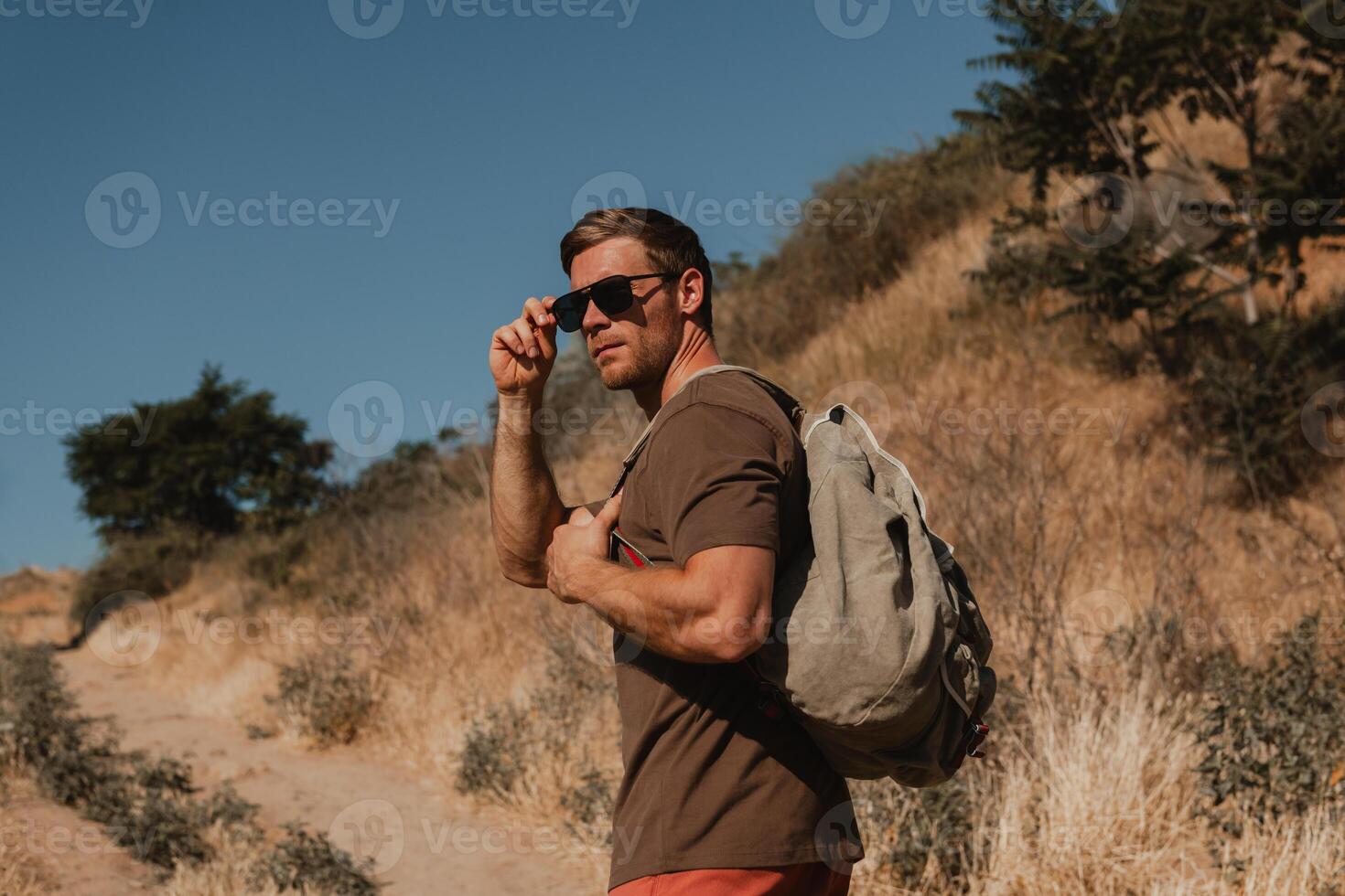 Confident male adventurer with backpack adjusting glasses while walking by the mountain footpath photo