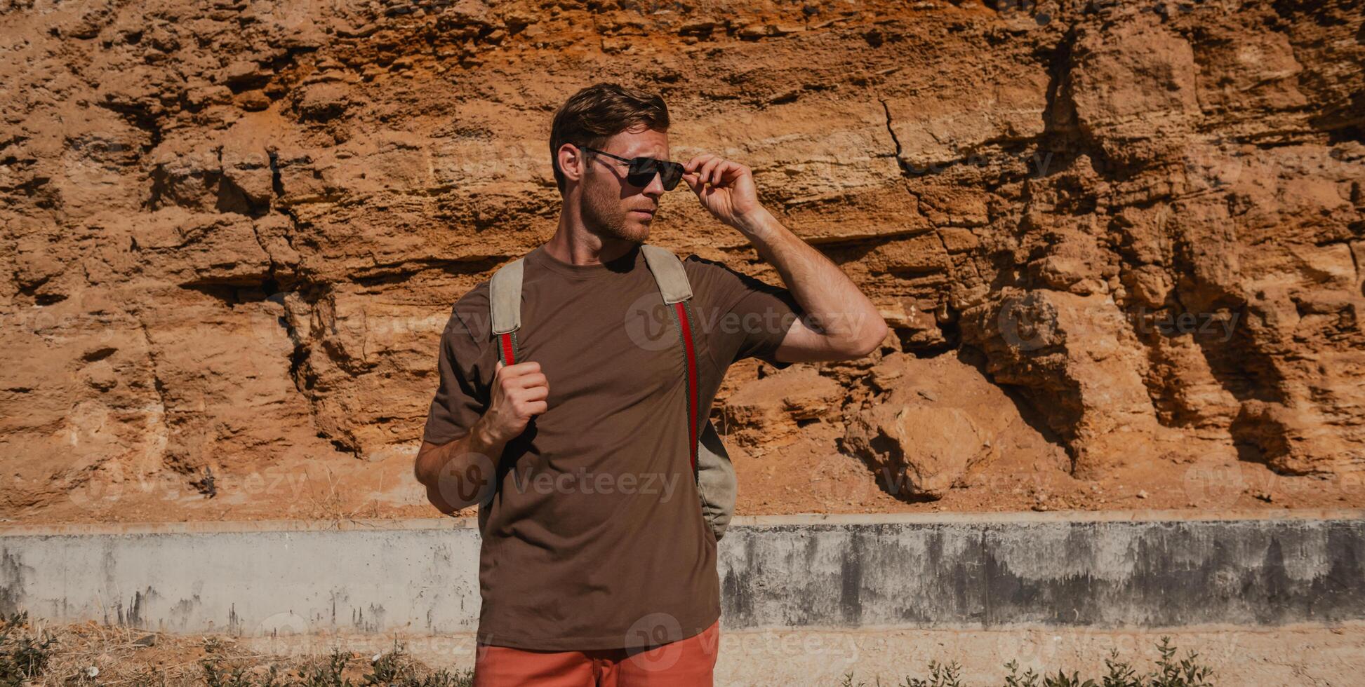 Confident adventurer with backpack adjusting glasses while standing against the rocky mountain photo