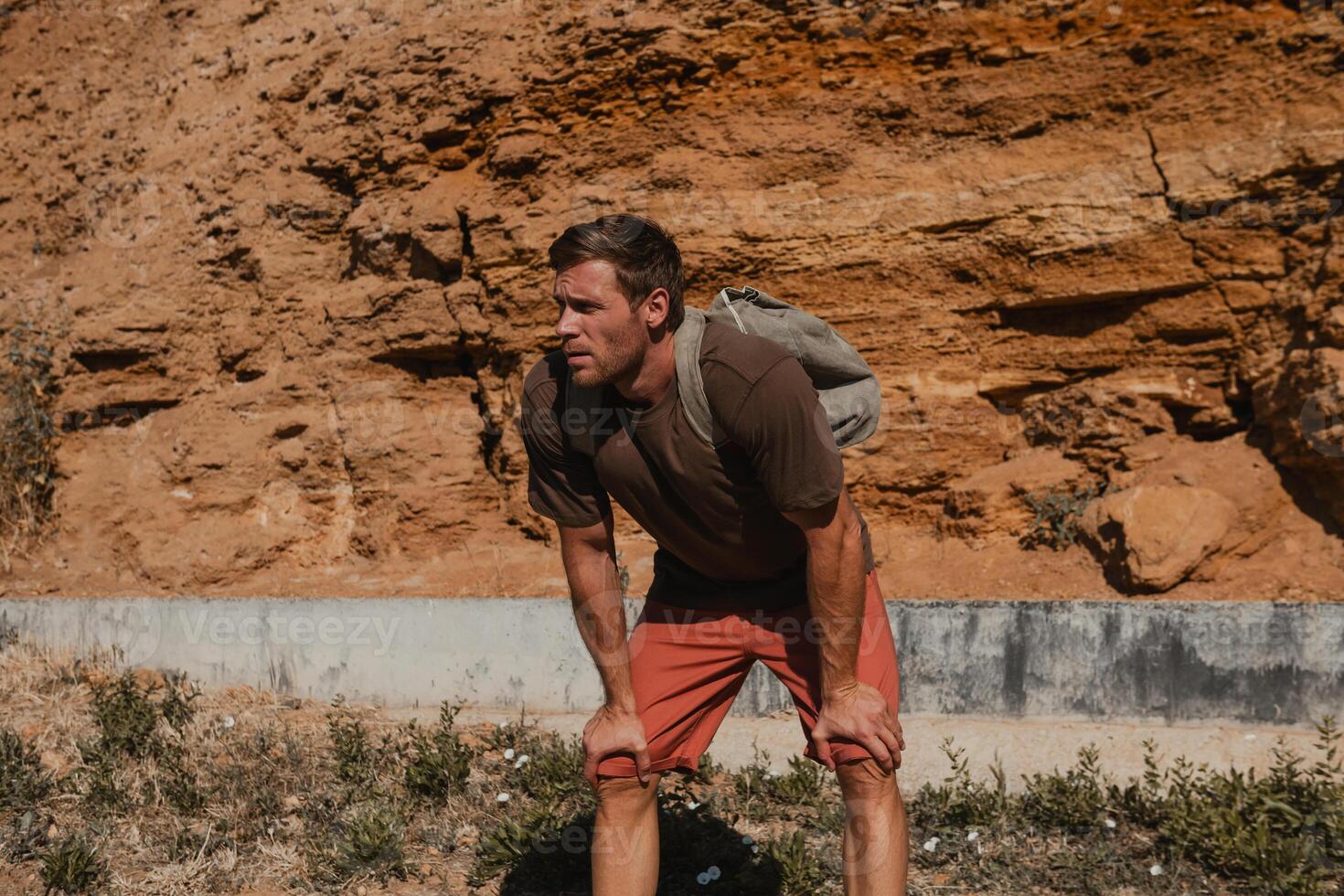 Tired adventurer with backpack holding hands on knees while resting against a rocky mountain photo