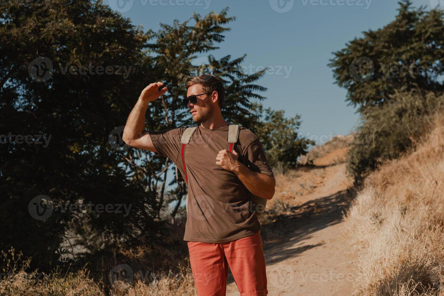 Confident adventurer with backpack covering face from sun while standing on the mountain footpath photo