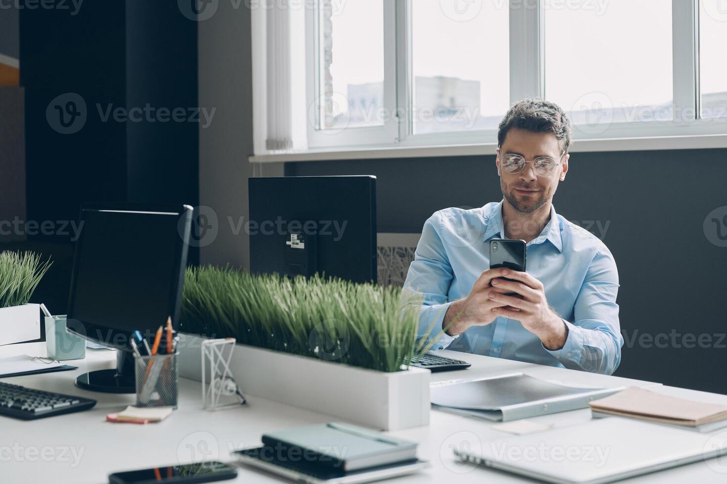 Confident young businessman using smart phone while sitting at his working place in office photo
