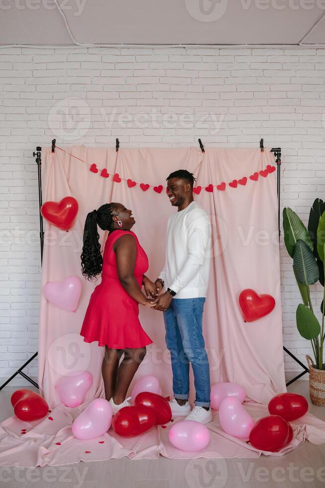 Happy young African couple holding hands while standing on pink fabric background with heart shaped balloons photo