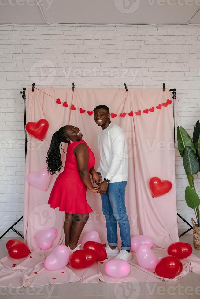 Smiling young African couple holding hands while standing on pink fabric background with heart shaped balloons photo