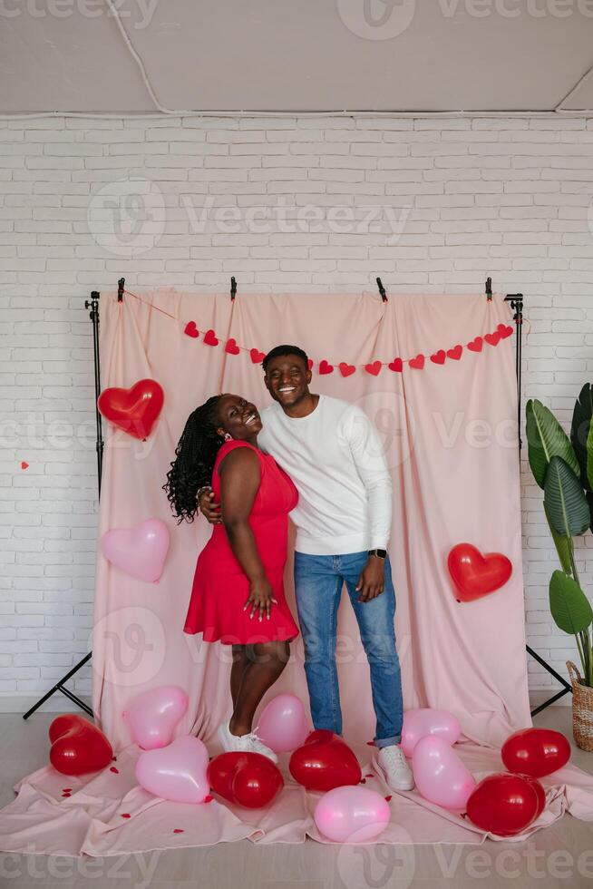 Happy young African couple embracing while standing on pink fabric background with heart shaped balloons photo
